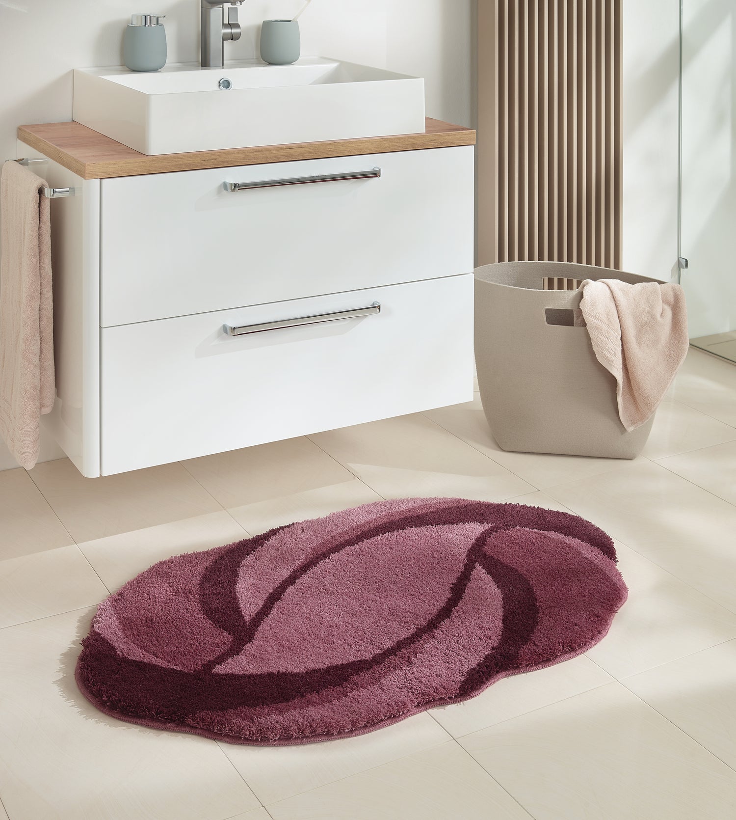 Ovaler Badteppich Kleine Wolke Creativo Valletta in Beere vor weißem Waschbecken im Bad.