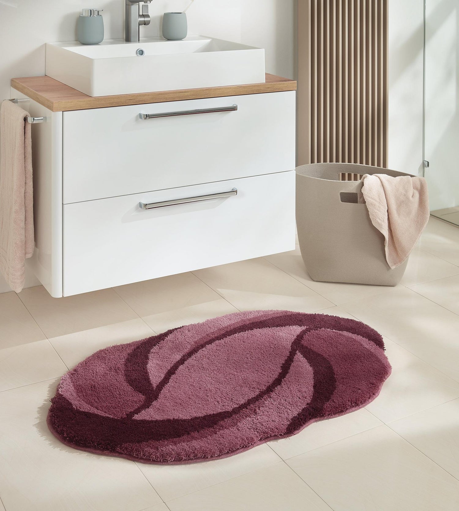 Ovale Badematte in Rosa- und Bordeaux-Tönen vor weißem Waschschrank