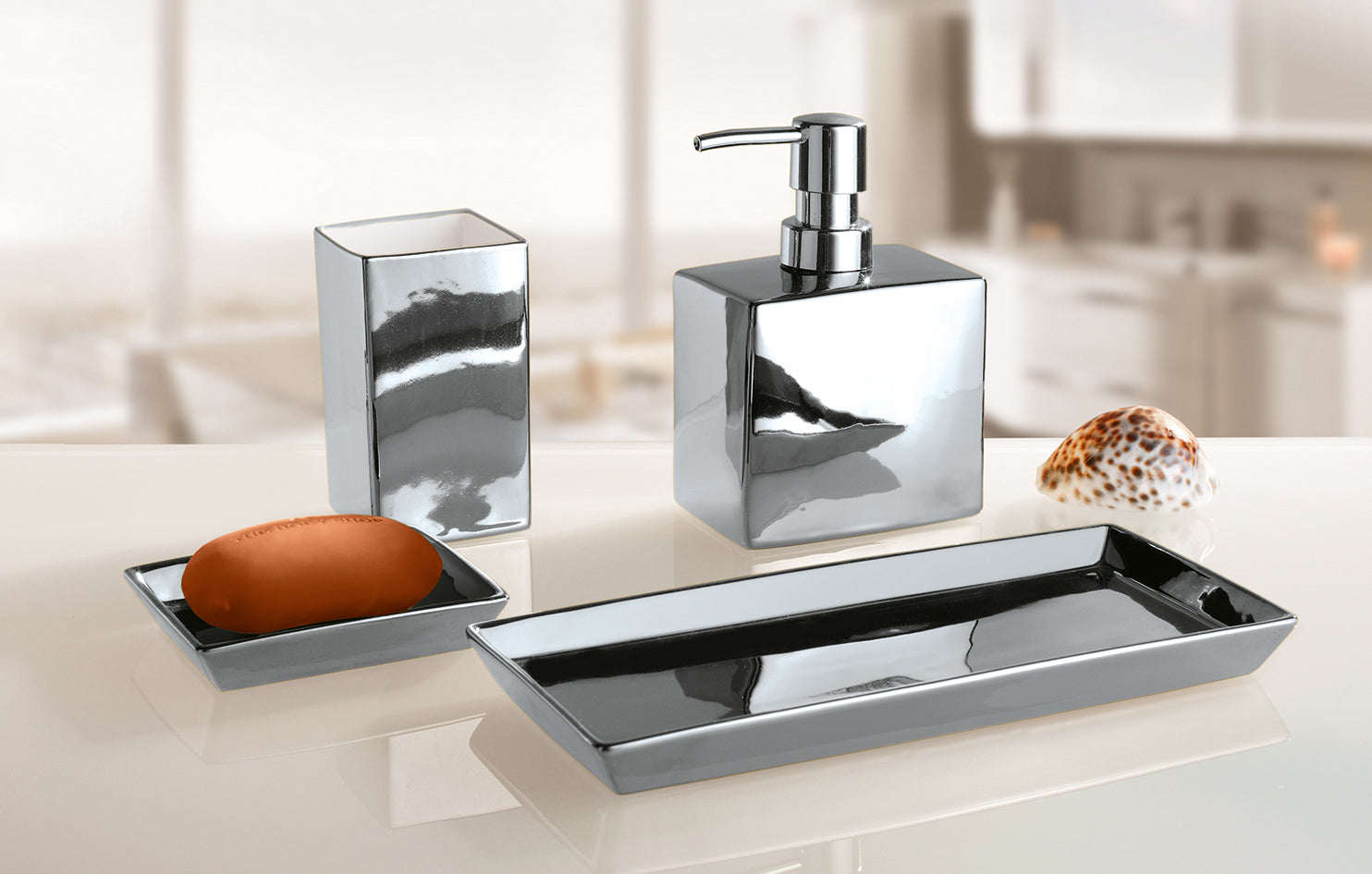 Silberfarbener Zahnputzbecher GLAMOUR steht mit Seifenspender und Schalen auf einem Waschbeckenrand.