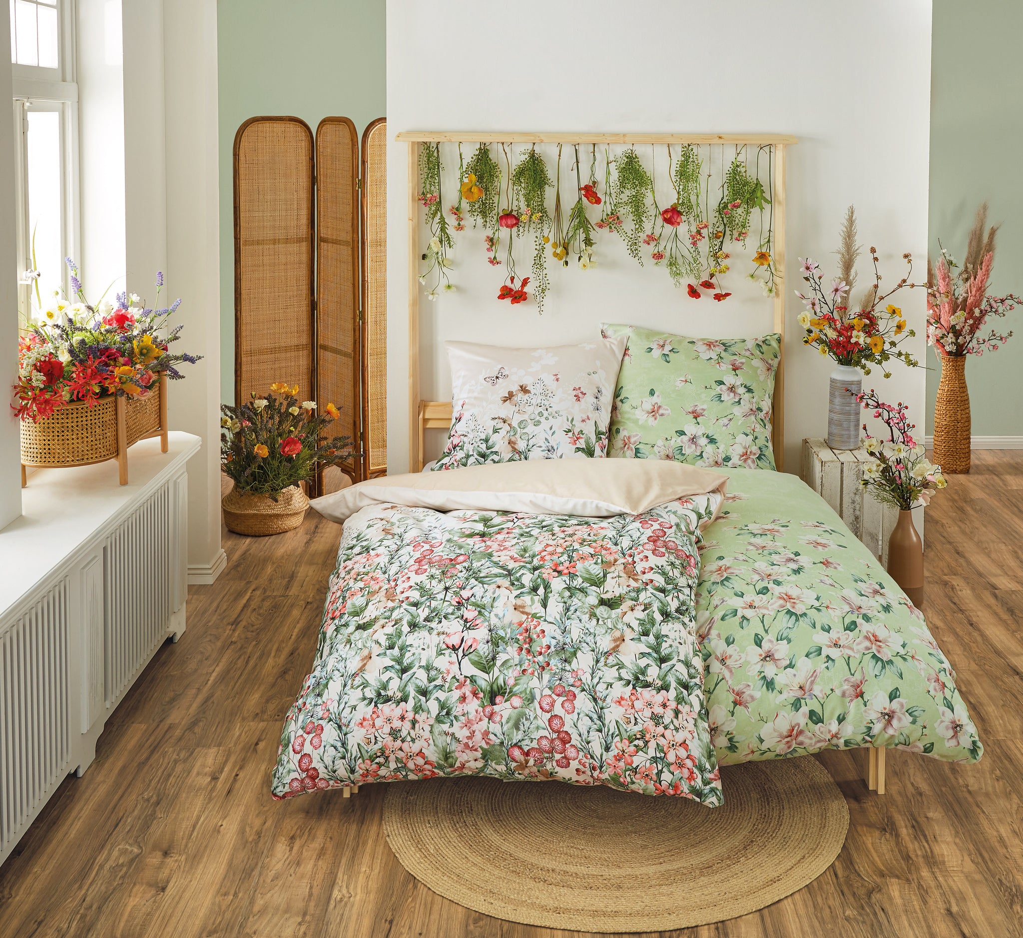 Bett mit geblümter Bettwäsche, Pflanzen und Blumen in hellem Raum