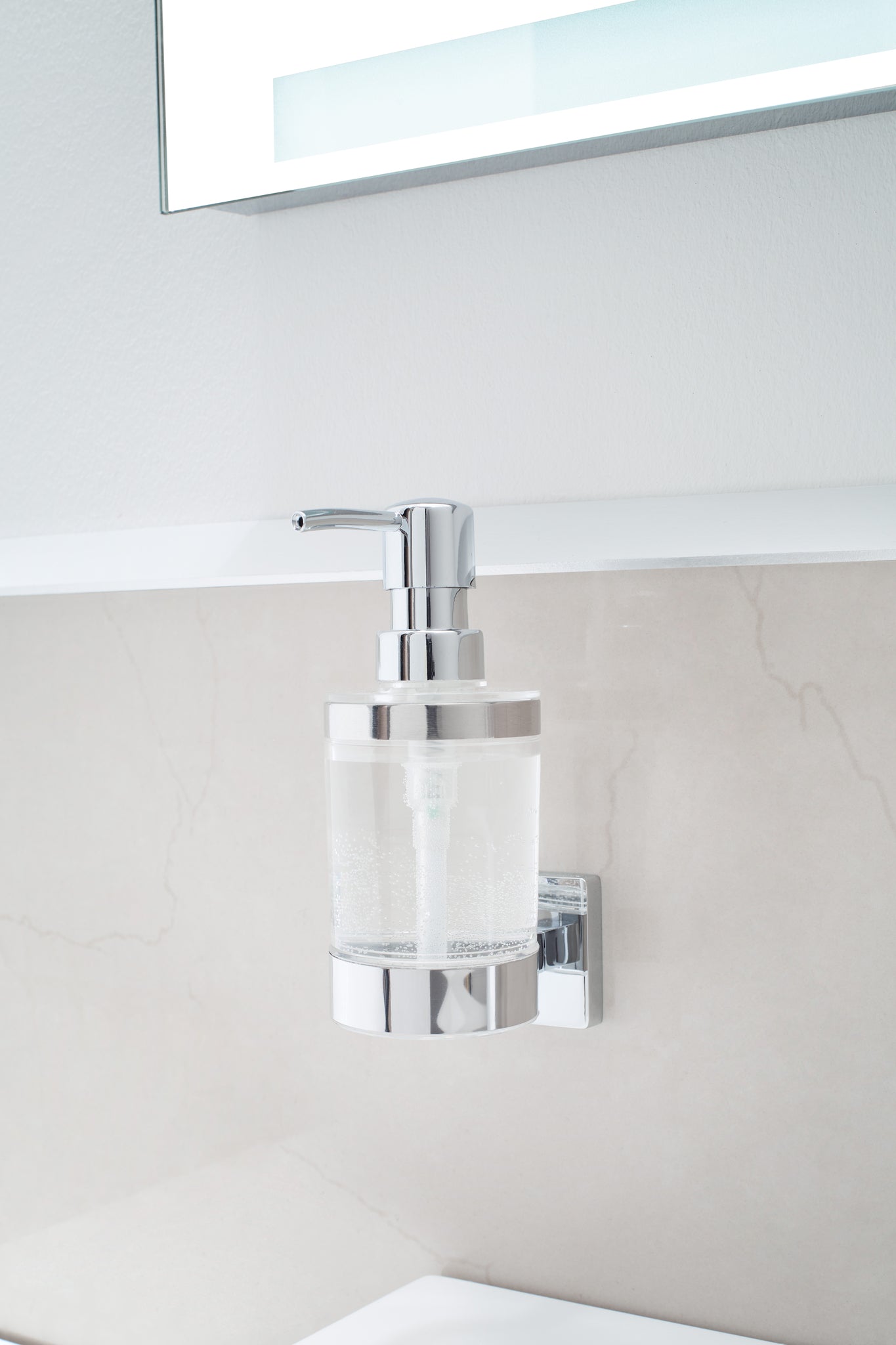 Wand-Seifenspender LUNO Clear aus Glas und Chrom, gefüllt, montiert an heller Badwand unter Spiegel.