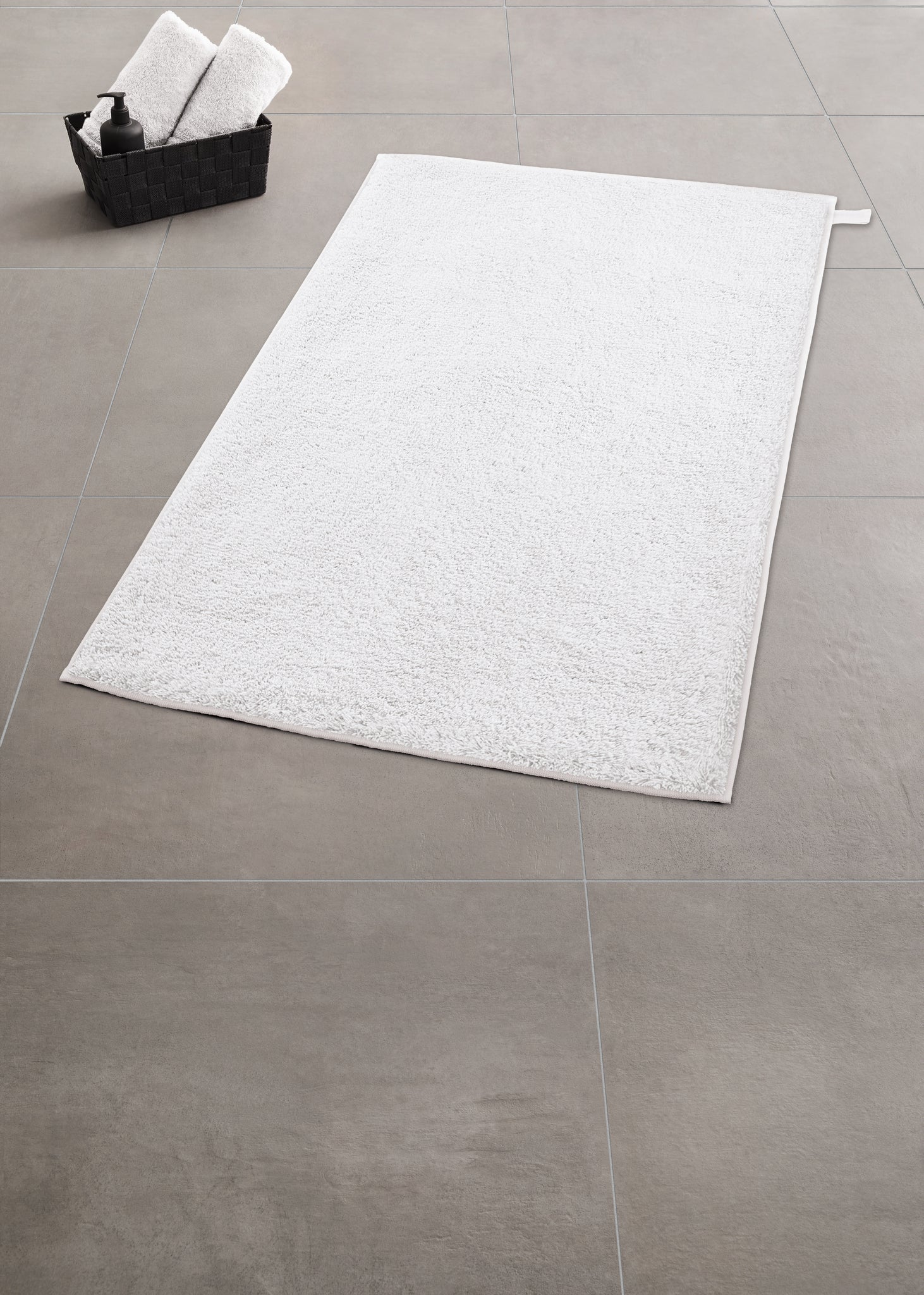 Weißer Badteppich VITA 50 x 80 cm auf grauem Fliesenboden neben schwarzem Korb mit Handtüchern.