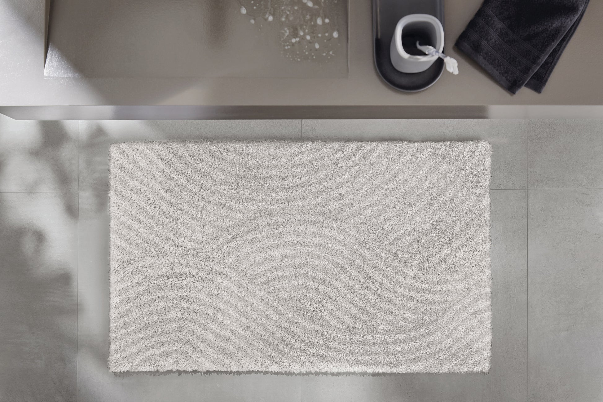 Badteppich CALMA Nebel 70 x 120 cm liegt vor einem Waschbecken auf grauem Badezimmerboden.