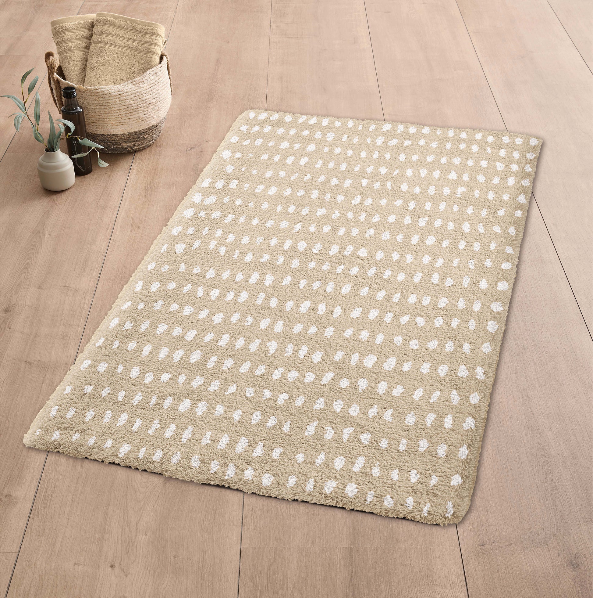 Badteppich DOTTINO Macadamia 60 x 90 cm liegt auf Holzfußboden neben Korb mit Handtüchern.