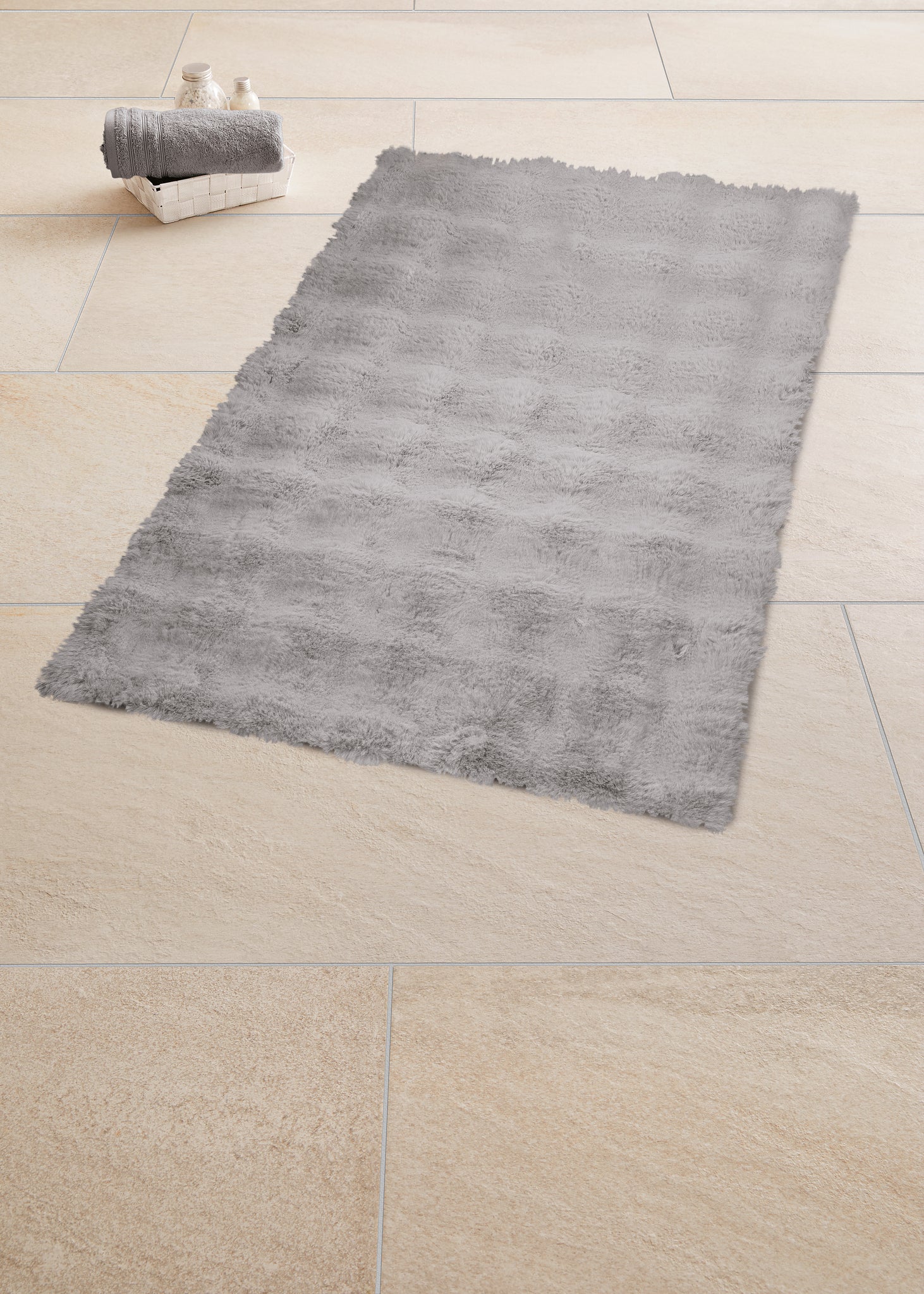 Badteppich CLOUD in Silbergrau 60 x 90 cm liegt auf beigen Fliesen neben einem Korb mit Handtuch.