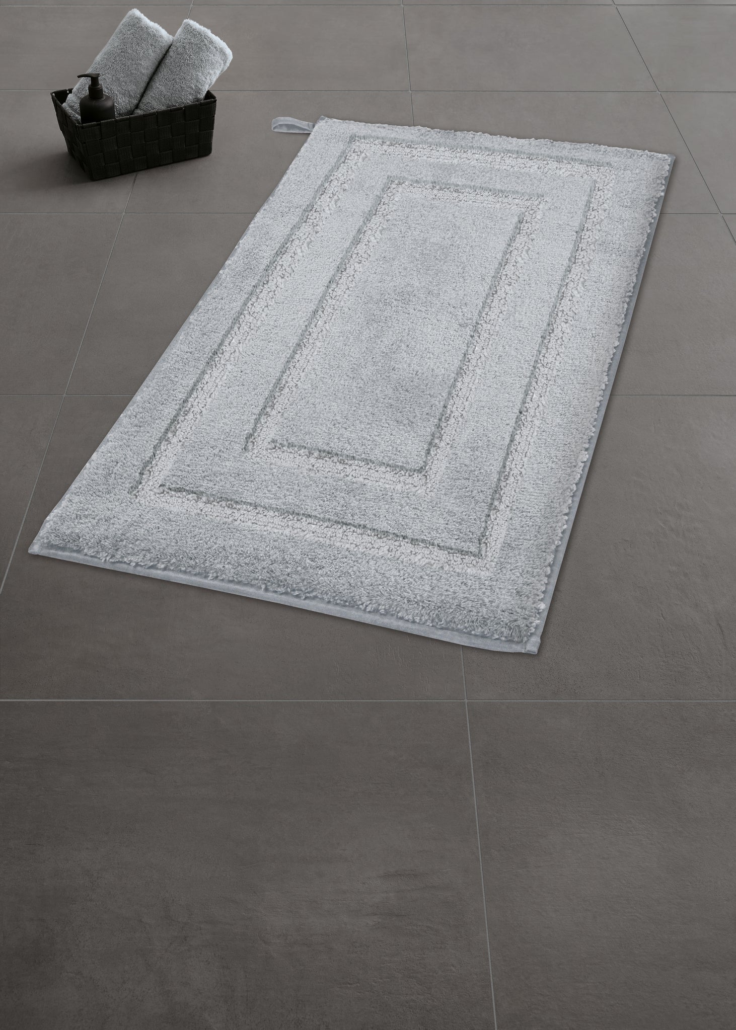Badteppich NICO Silbergrau 50 x 80 cm liegt auf grauen Fliesen neben einem Korb mit Handtüchern.