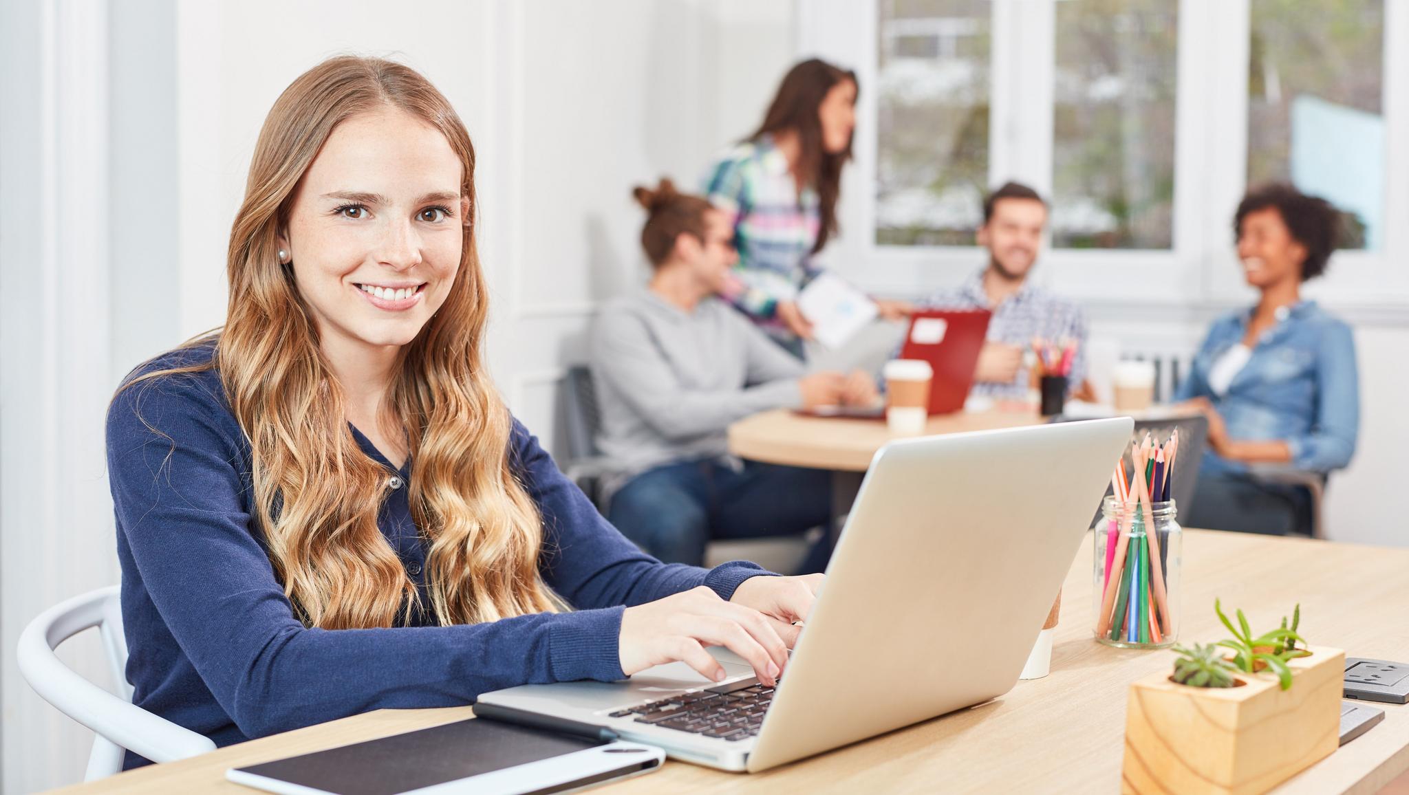 Junge Frau arbeitet lächelnd am Laptop, Kollegen im Hintergrund Büro
