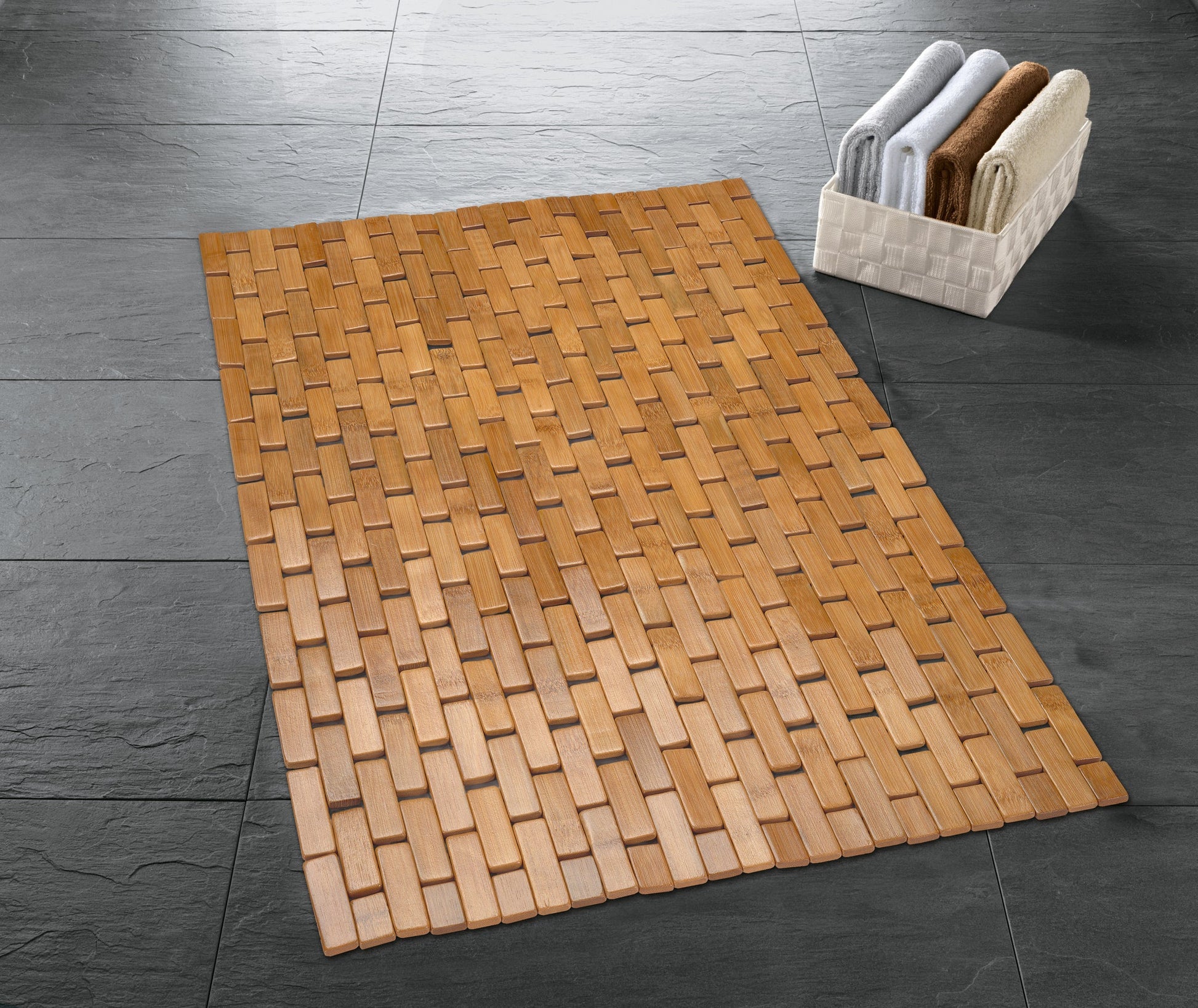 Badematte aus Bambus PALITO Natur 50 x 70 cm liegt auf dunklen Fliesen neben einem Korb mit Handtüchern.