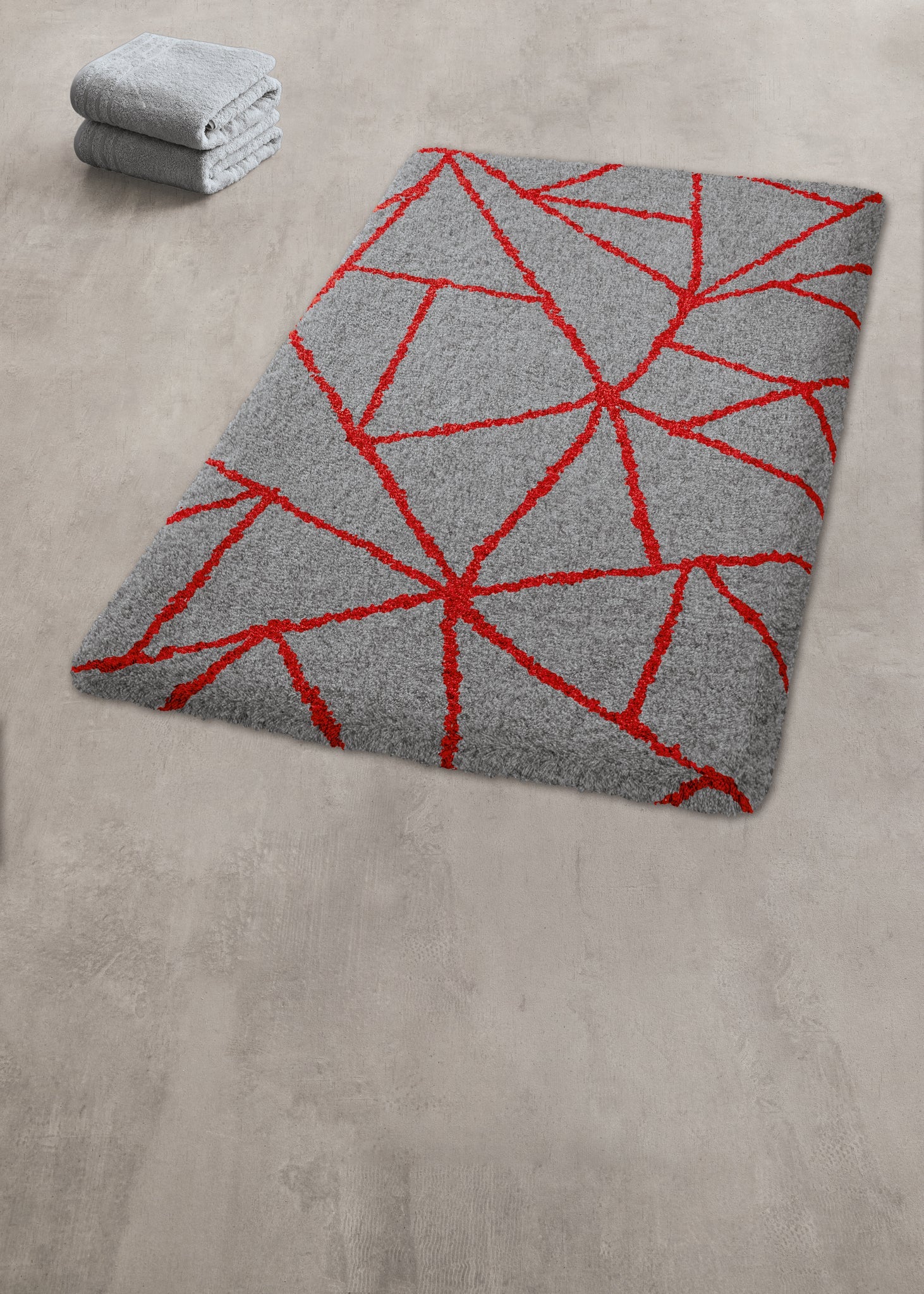 Badteppich BONDI Rubin 60 x 90 cm liegt graurot gemustert im Bad neben gefalteten Handtüchern.