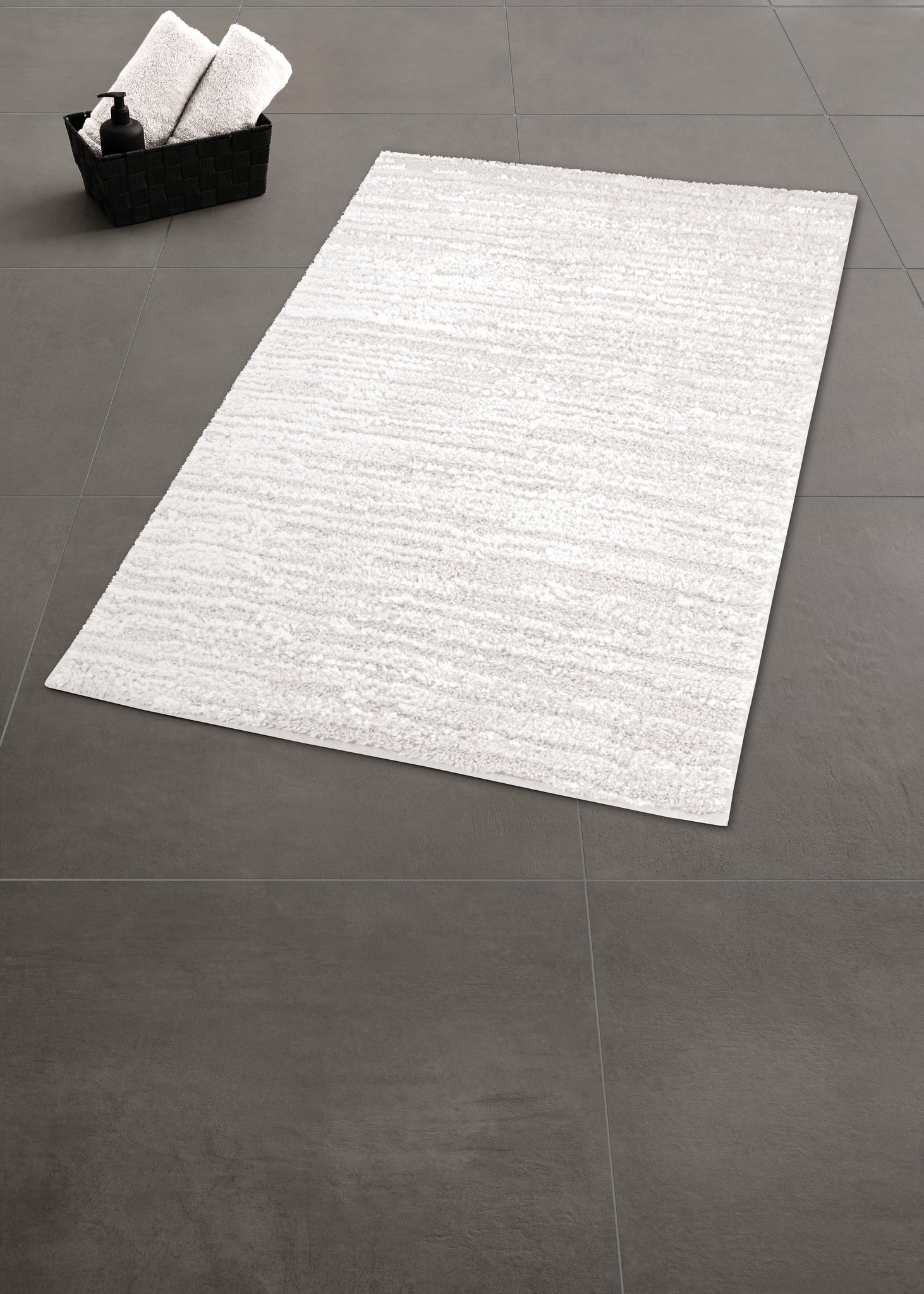 Hellgraue Badematte auf dunklem Fliesenboden mit Korb im Hintergrund