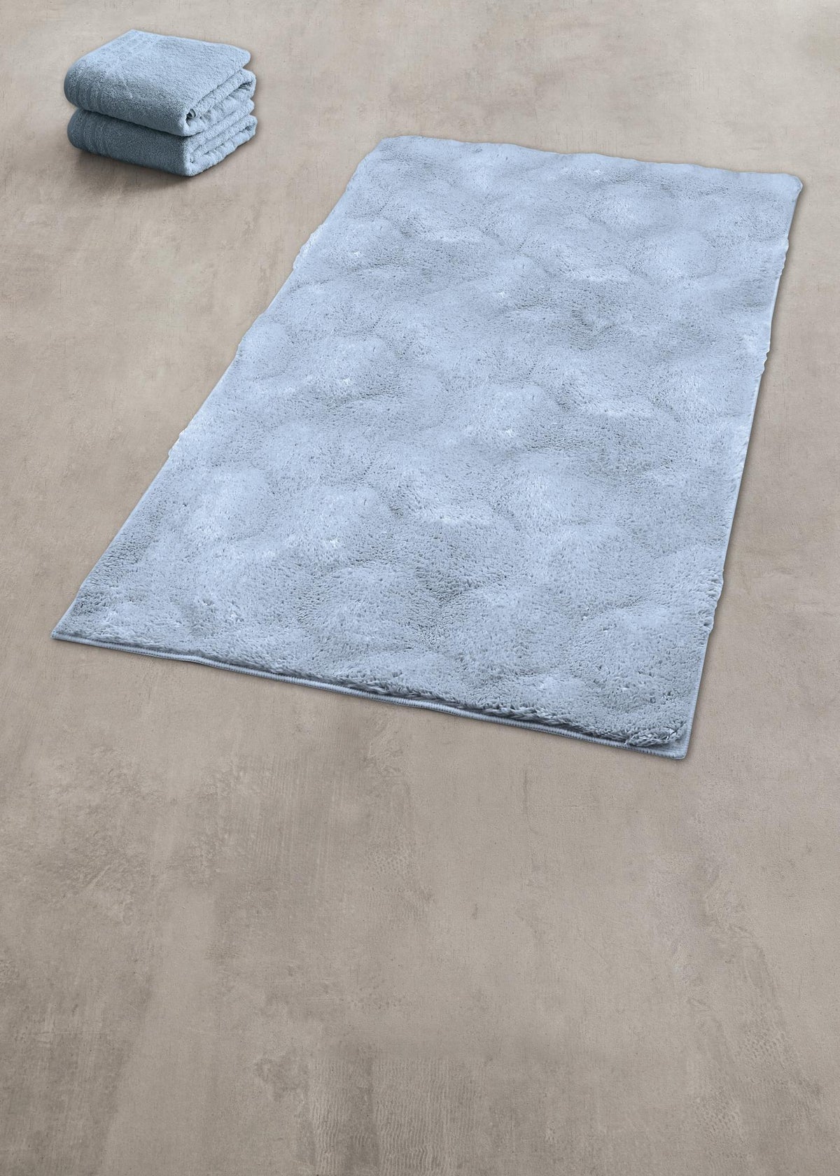 Hellblauer flauschiger Badteppich auf beigem Boden neben Handtüchern.