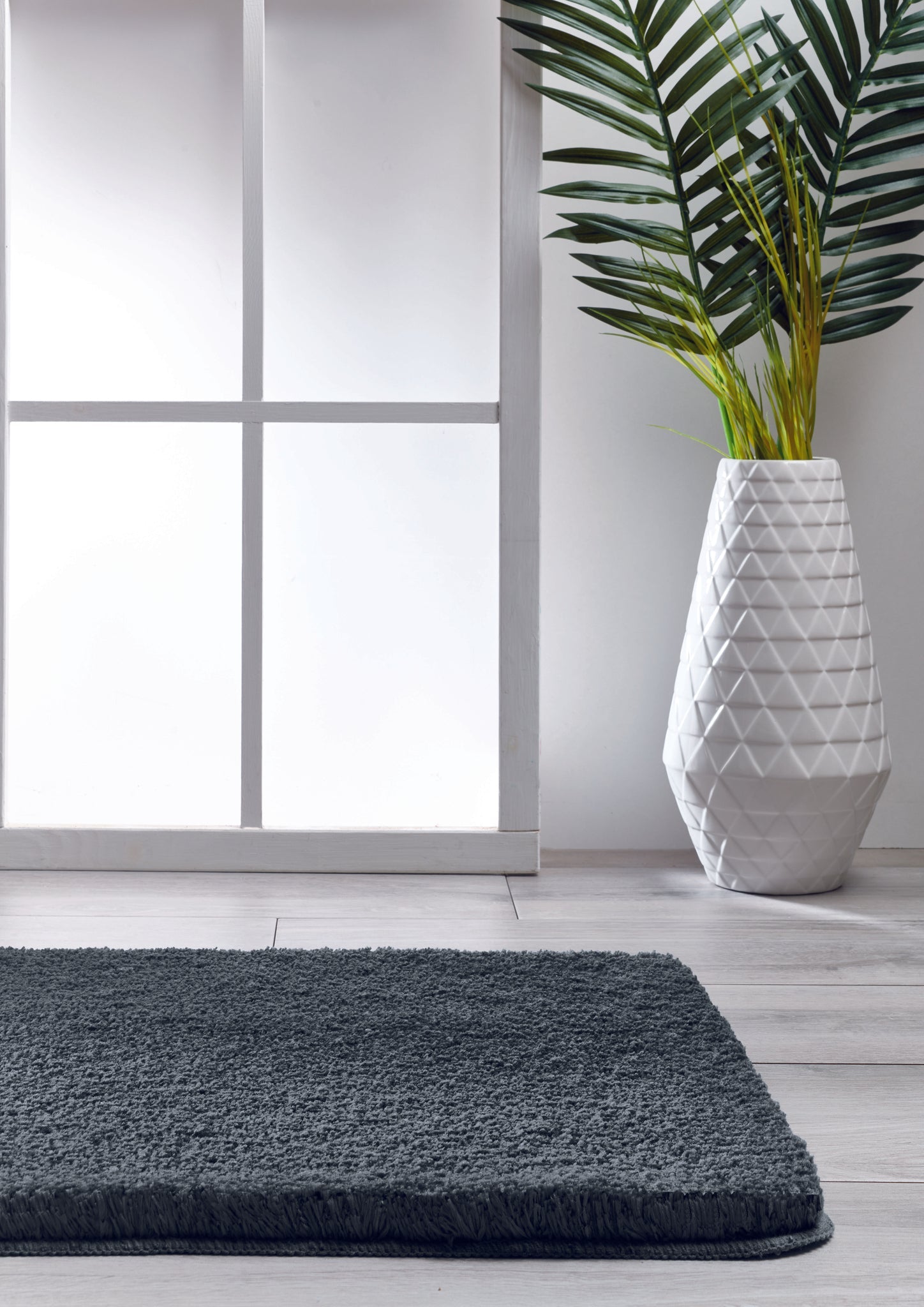 Dunkelgraue Badematte vor Fenster, daneben weiße Vase mit großer Pflanze
