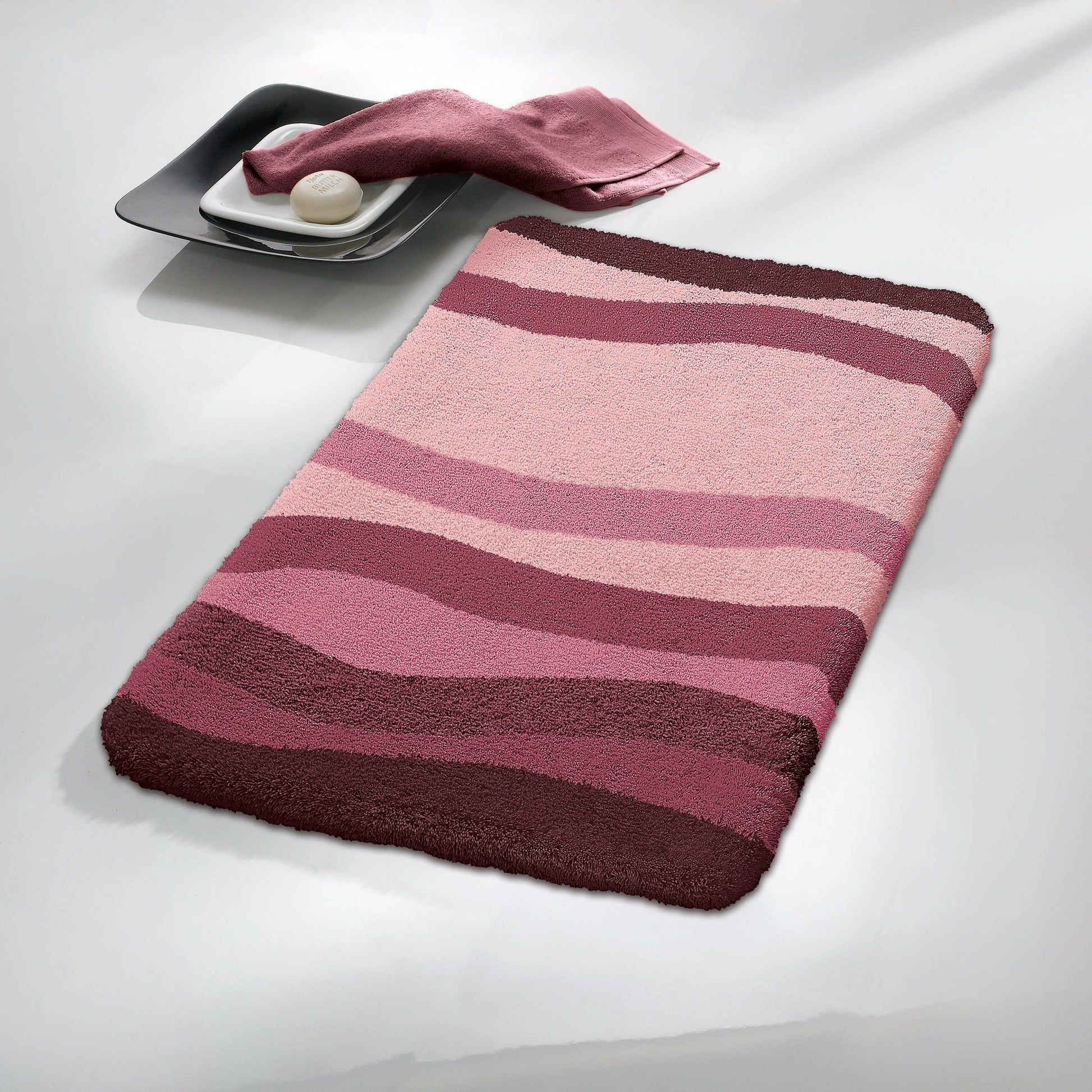 Badteppich MIAMI Beere 65 x 115 cm mit welligen Beere-Streifen neben Schale mit Seife und Tuch.