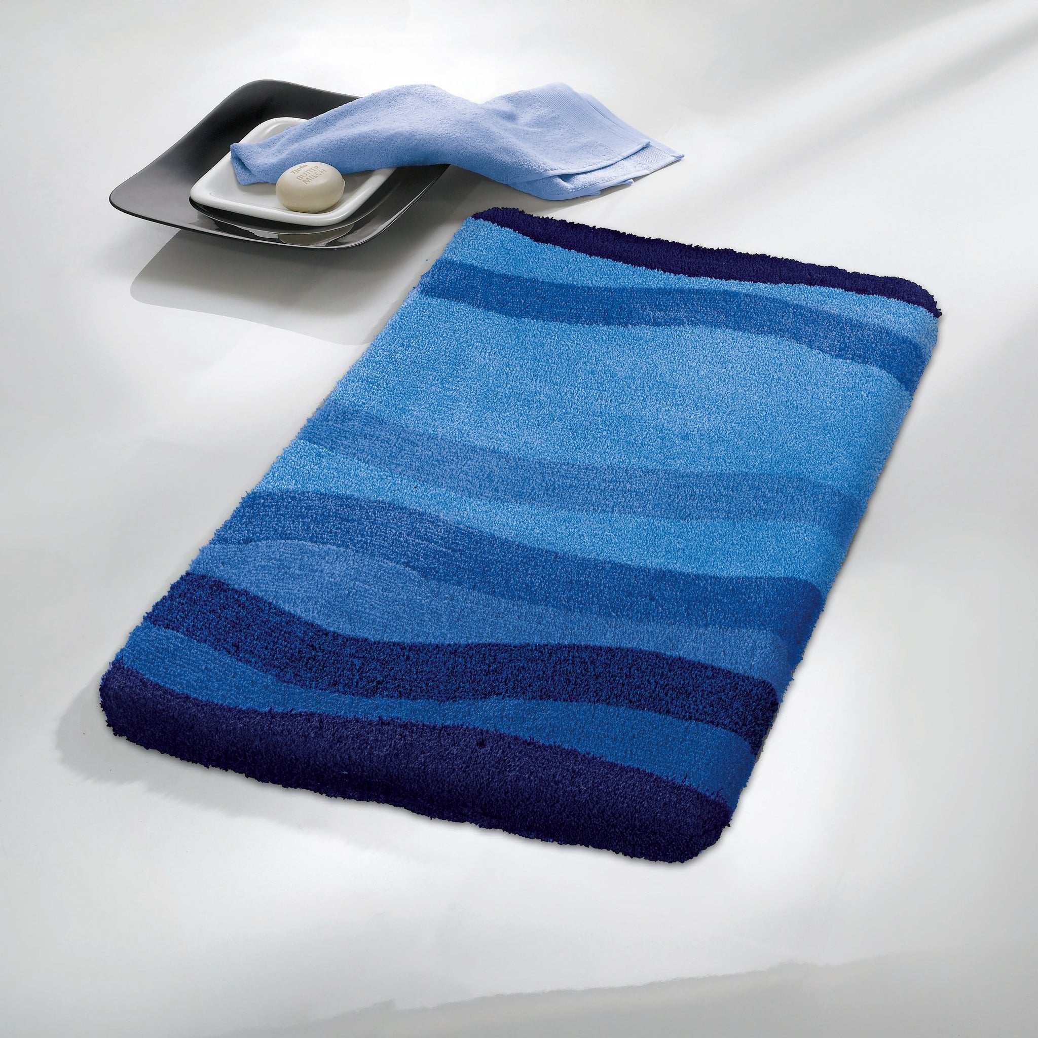 Badteppich MIAMI Himmelblau 60 x 90 cm liegt auf hellem Boden neben Seife und Handtuch.