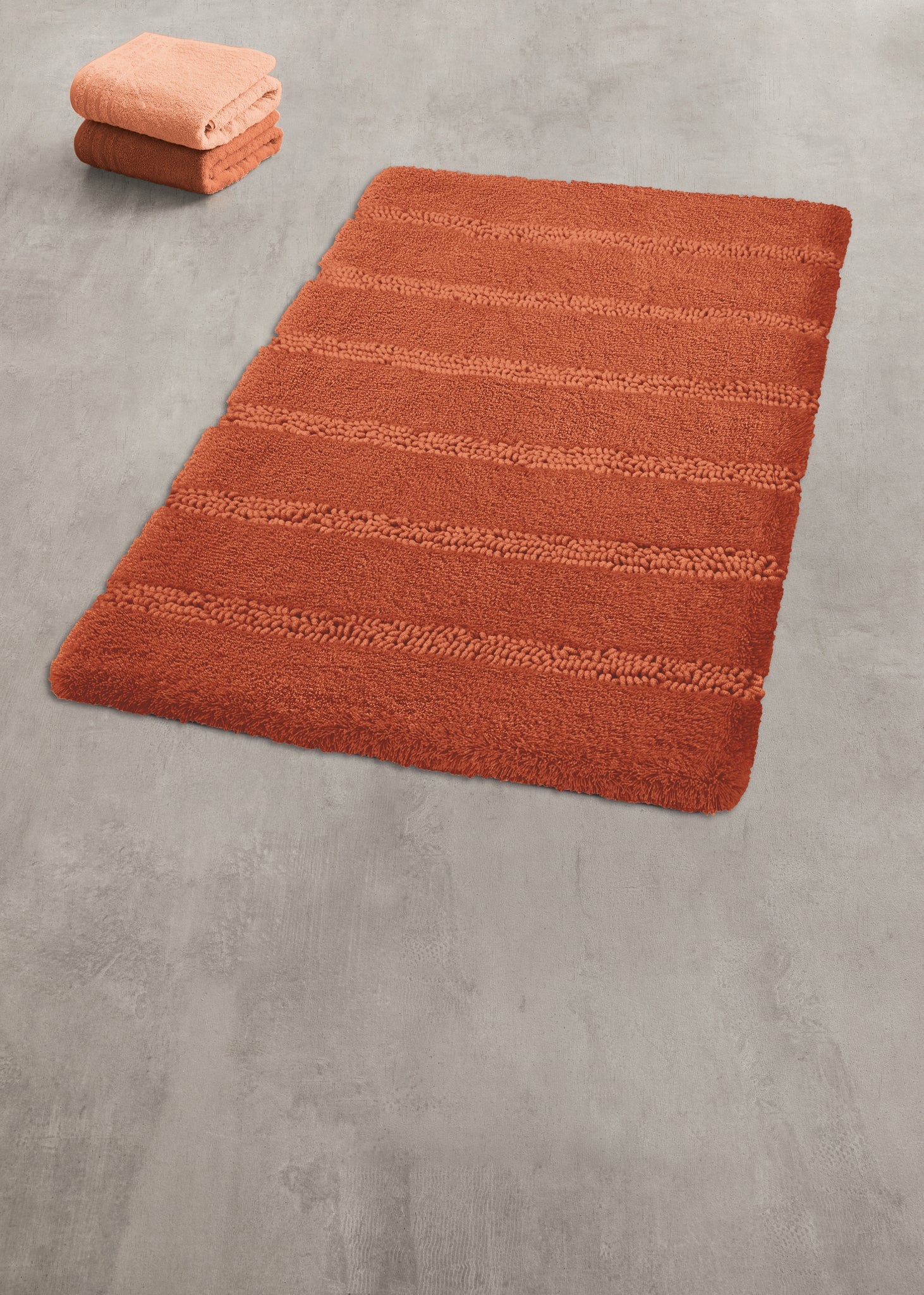 Oranger, rechteckiger Badteppich auf grauem Boden, gefaltete Handtücher
