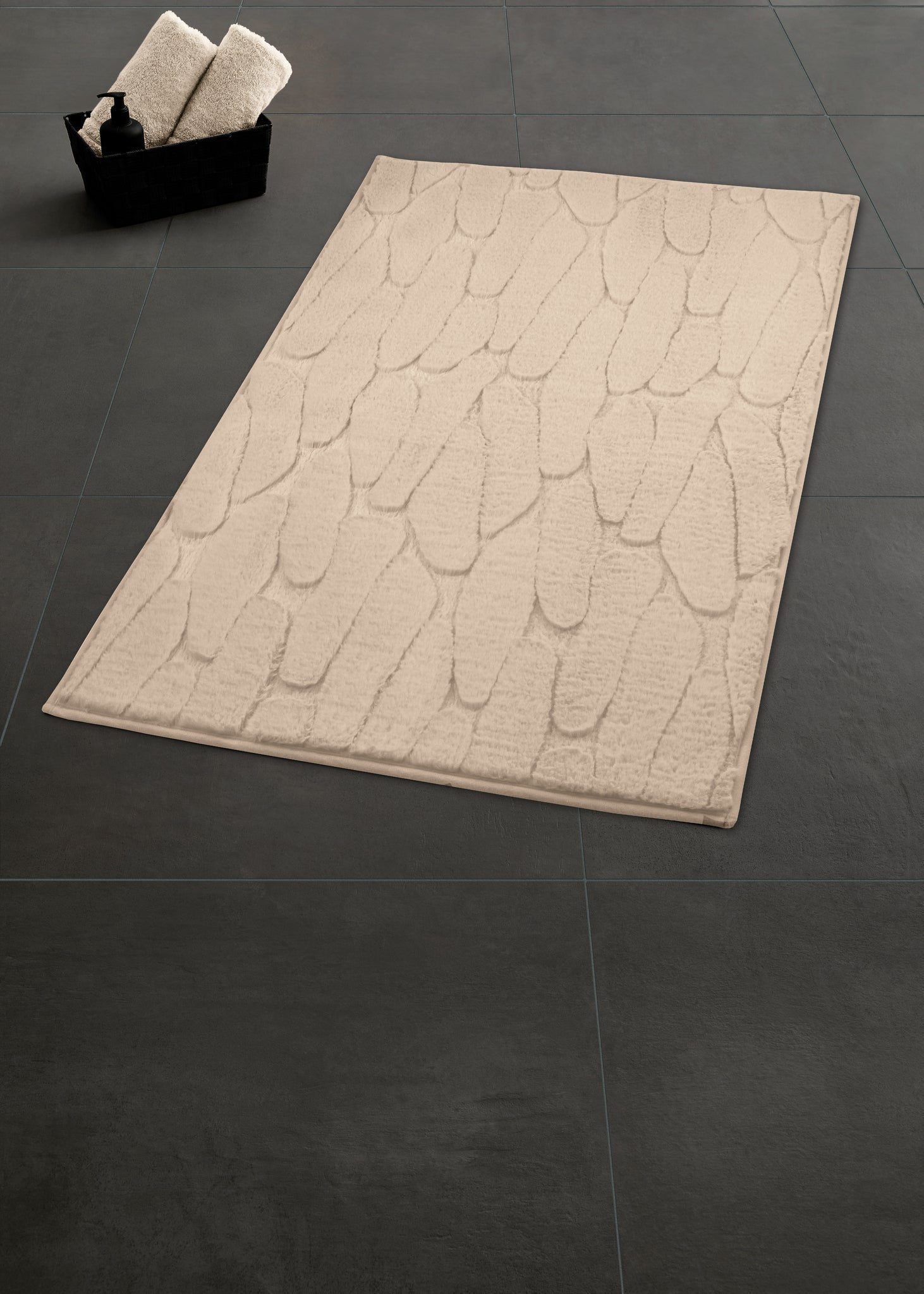 Badteppich TONDO seidenfarben 60 x 90 cm liegt auf dunklem Fliesenboden neben einem Korb mit Handtüchern.