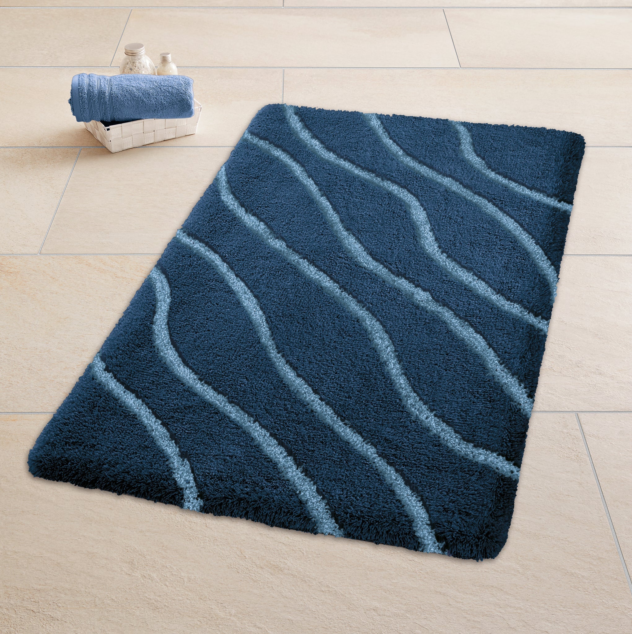 Badteppich TRAM Mare 55 x 65 cm liegt auf hellen Fliesen neben einem Korb mit Handtüchern.
