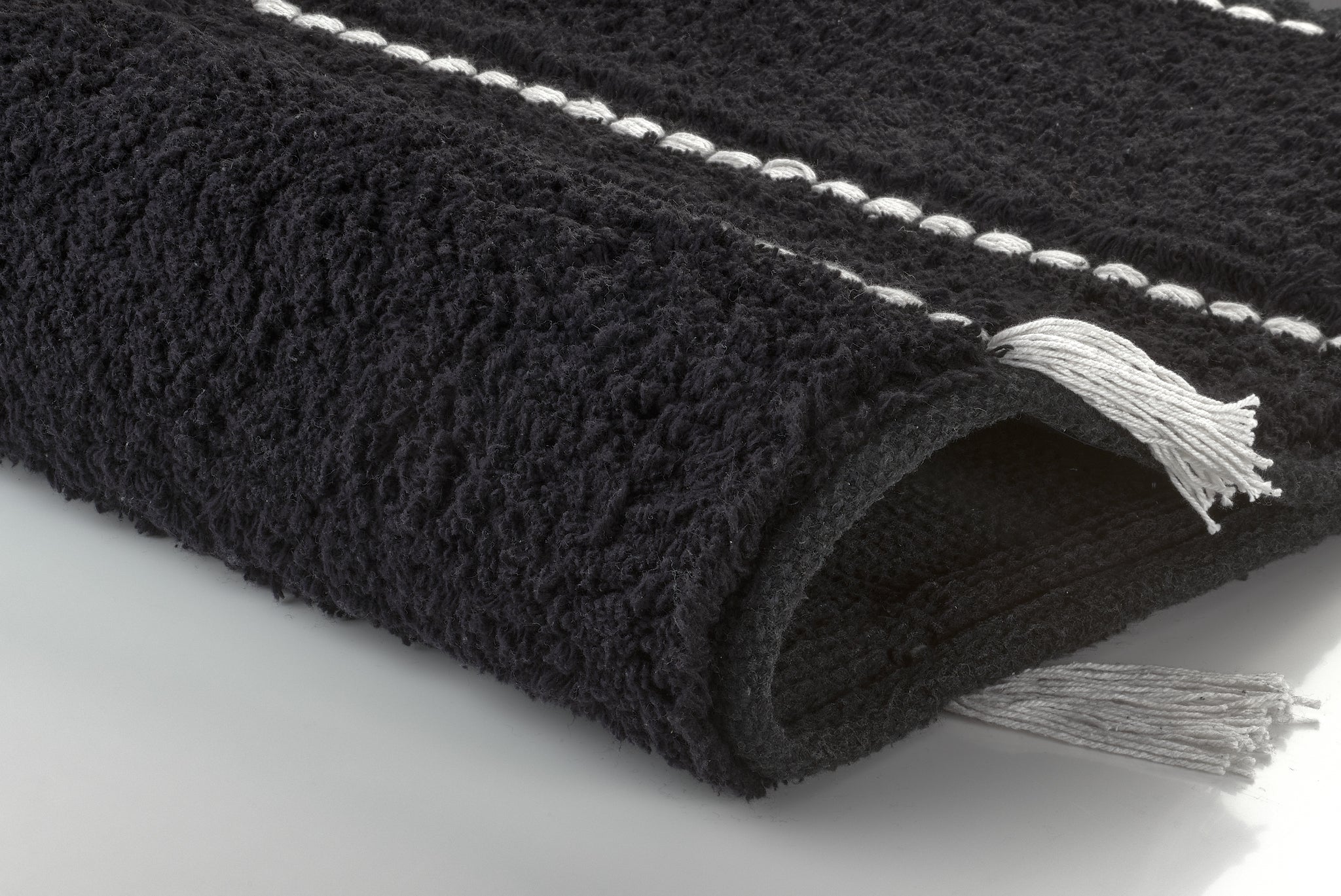 Badteppich TRAVIS Schwarz 55 x 65 cm liegt gerollt mit Fransen und weißen Streifen auf hellem Untergrund.