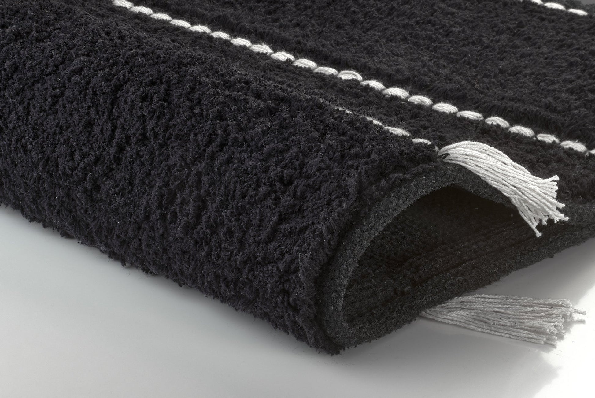 Schwarzer Badteppich TRAVIS mit strukturierter Oberfläche und hellen Fransen liegt auf hellem Boden.