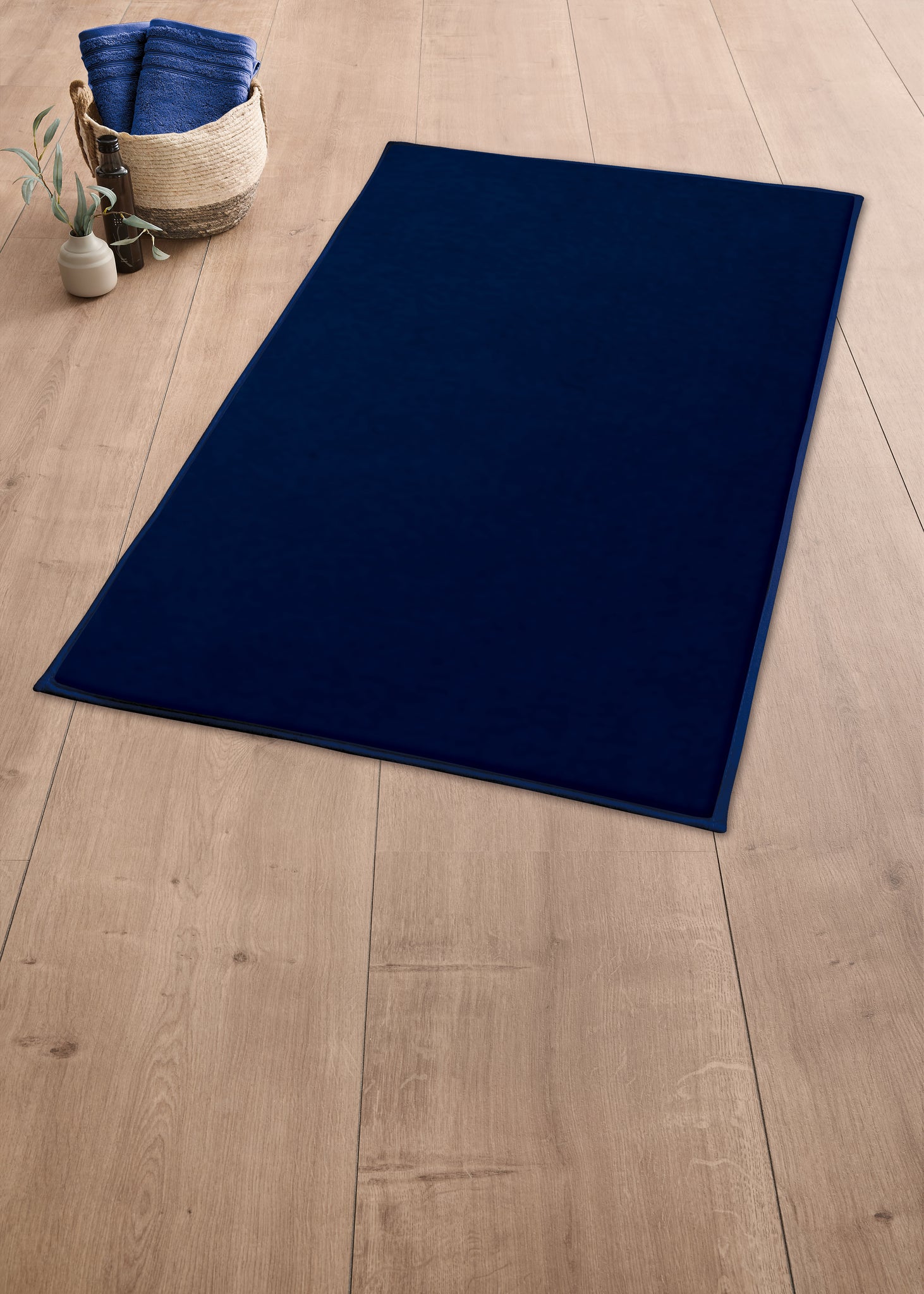 Badteppich VELVET Navy 60 x 90 cm liegt auf Holzfußboden neben Korb mit Handtüchern und Pflanze.