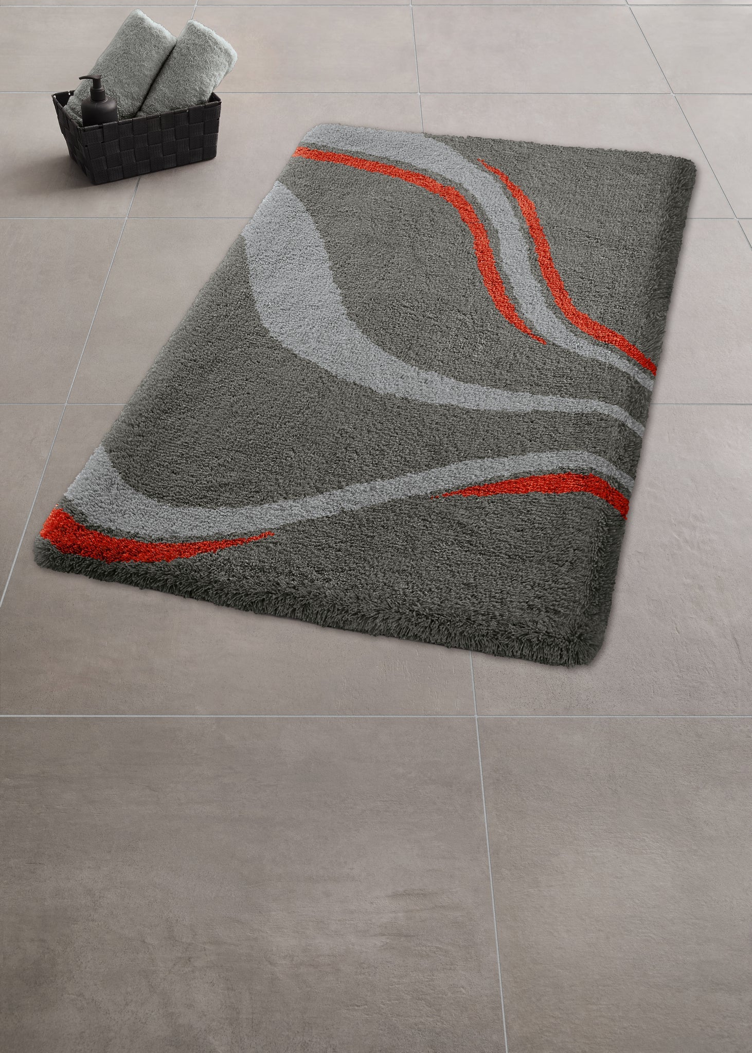 Badteppich VENTO Schiefer 60 x 90 cm liegt auf grauen Fliesen neben einem Korb mit Handtüchern.