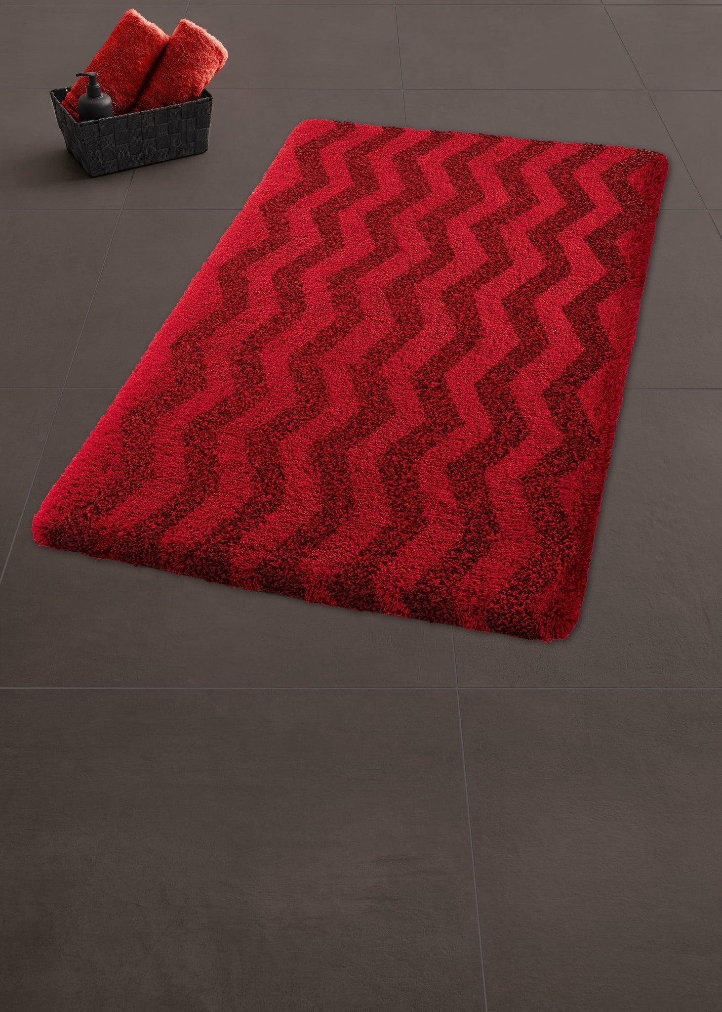 Badteppich ZACK Rubin 60 x 90 cm liegt auf dunklem Badezimmerboden neben Korb mit roten Handtüchern.