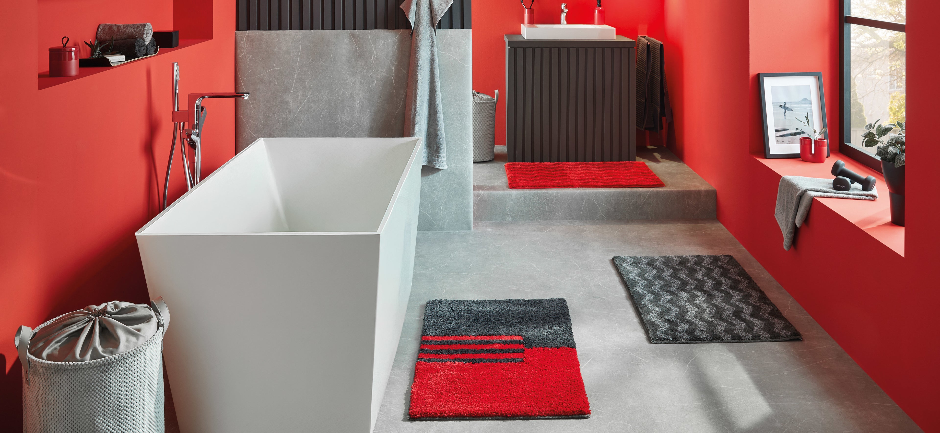 Moderne Badezimmer-Szene mit Badteppichen Dynamic Red vor roter Wand und weißer Badewanne.