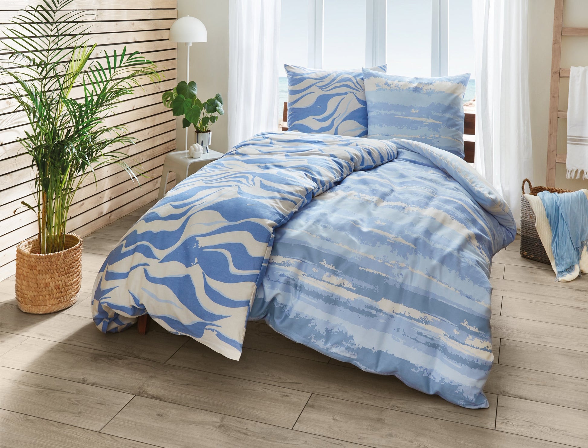 Bett mit blau-beiger Bettwäsche im Wellenmuster neben grüner Zimmerpflanze