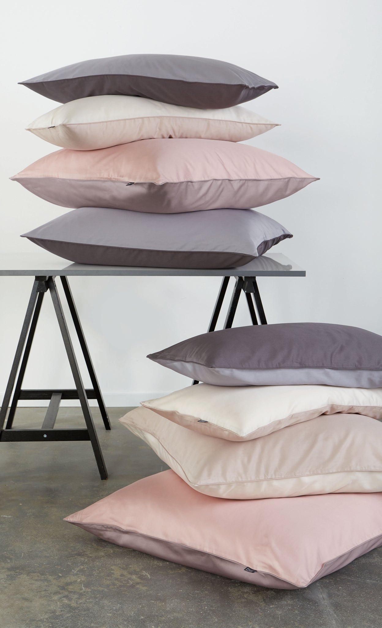 Mehrere Kissen der Bettwäsche Satin LINDA Old Rose 80x80 cm liegen gestapelt auf Tisch und Boden.