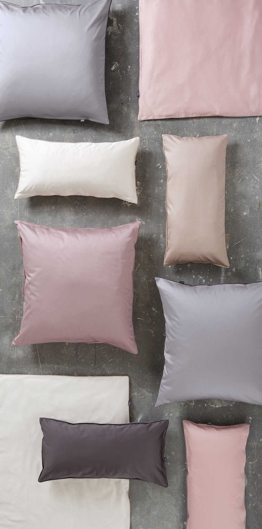 Mehrere Kissen und Decken aus Bettwäsche Satin LINDA Taupe in verschiedenen Größen liegen auf grauem Boden.