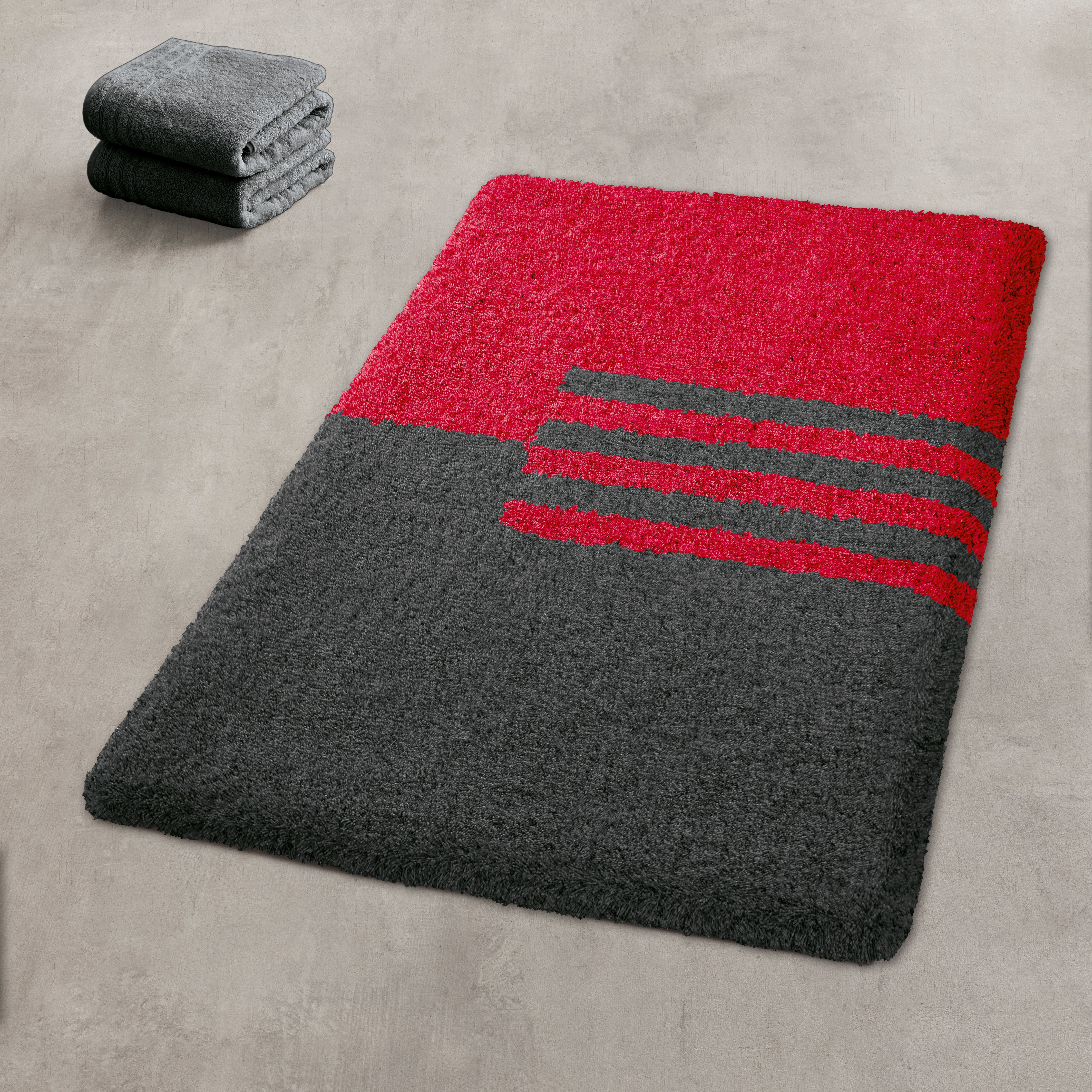 Roter und anthrazitfarbener Badteppich mit Streifen, daneben gefaltete passende Handtücher.