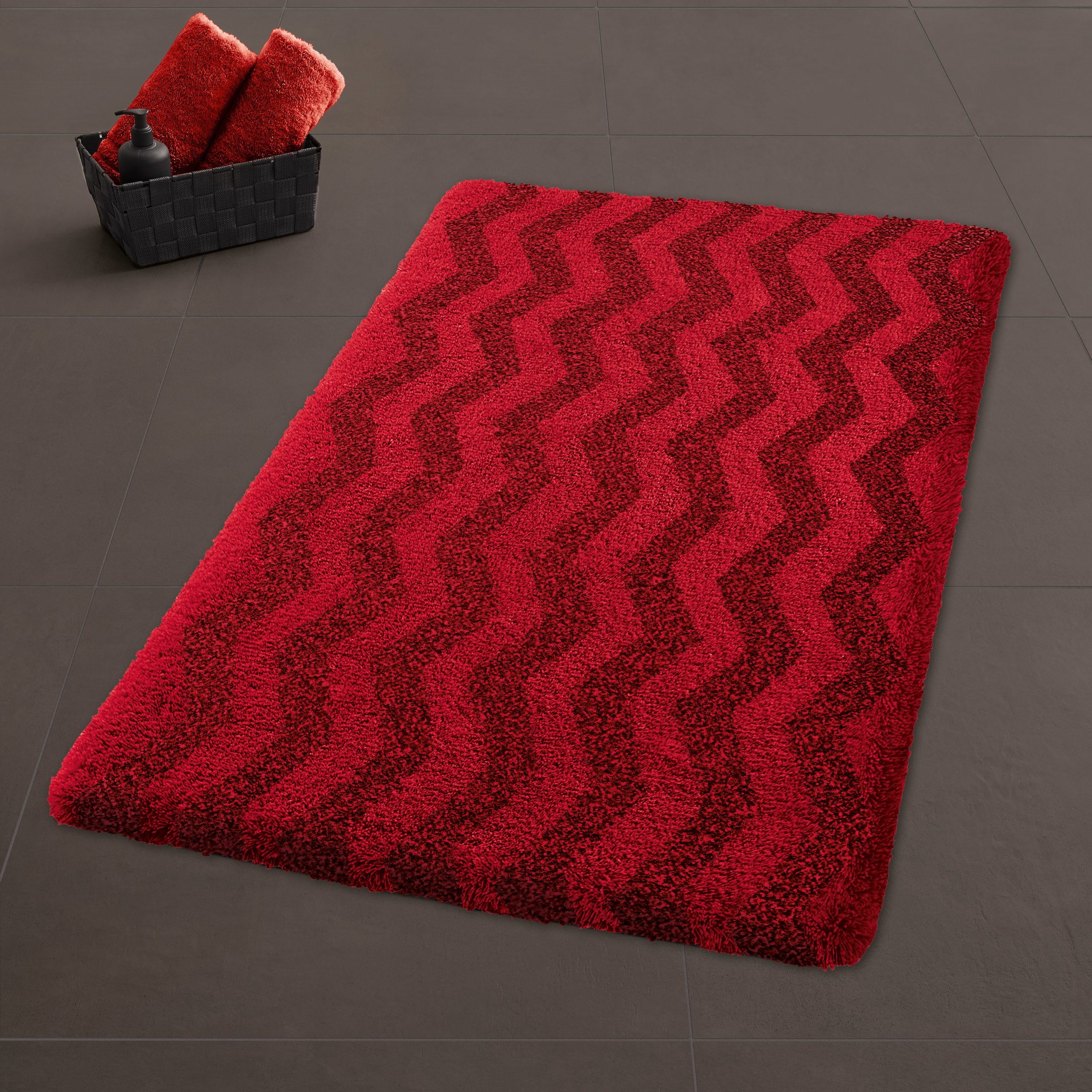 Roter Badteppich Kleine Wolke Dynamic Red mit Zickzackmuster auf dunklen Fliesen neben Korb mit Handtüchern.