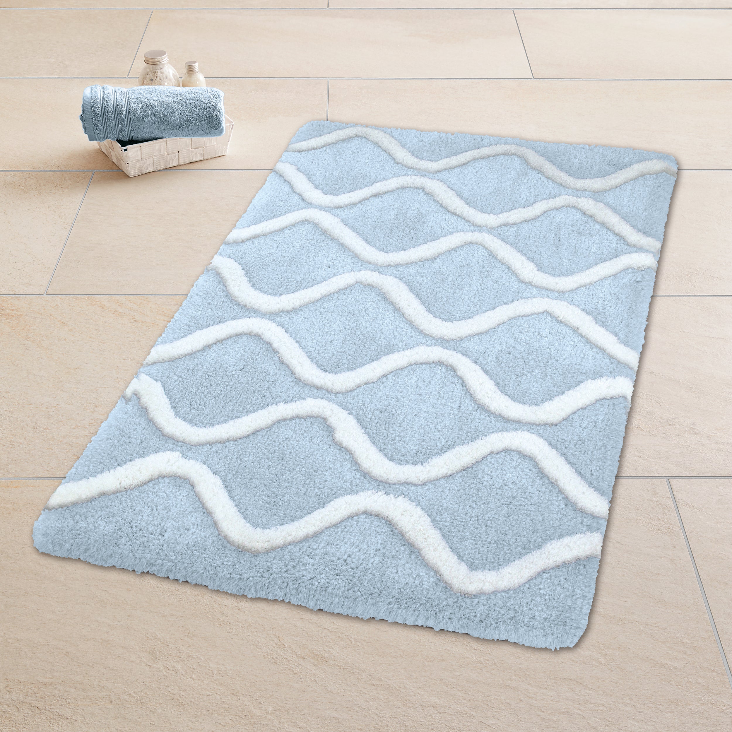 Badematte Kleine Wolke Piana in Hellblau mit weißen Wellen liegt auf beigem Fliesenboden.