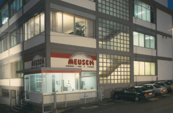 Abendliche Außenansicht eines Firmengebäudes mit Meusen-Logo, Glasfassade, Treppe und parkenden Autos.