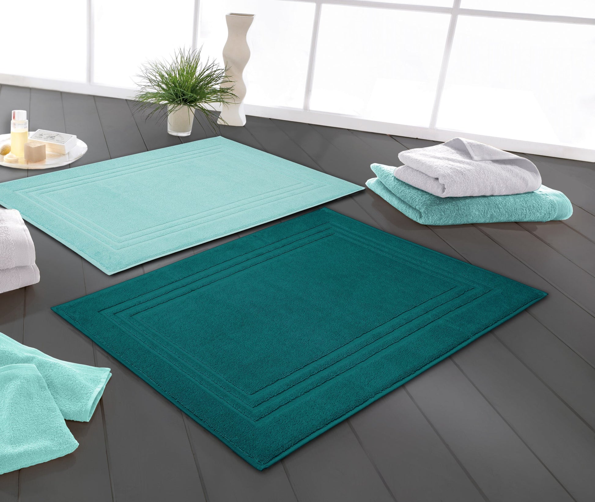 Zwei türkisfarbene Badematten auf dunklem Boden neben Handtüchern