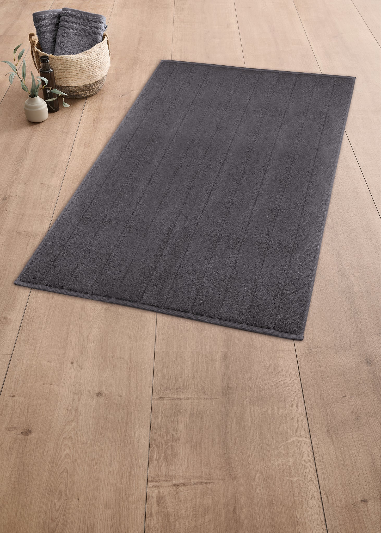 Graue rechteckige Badematte auf Holzfußboden neben Korb mit Handtüchern
