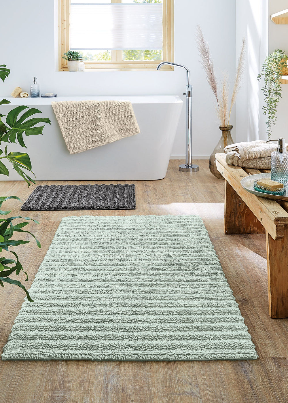 Helles Badezimmer mit Badewanne und Holzboden auf dem nachhaltig produzierte Badezimmerteppiche liegen. Holzbank mit Bad-Accessoires aus Glas und Frottier-Handtüchern.