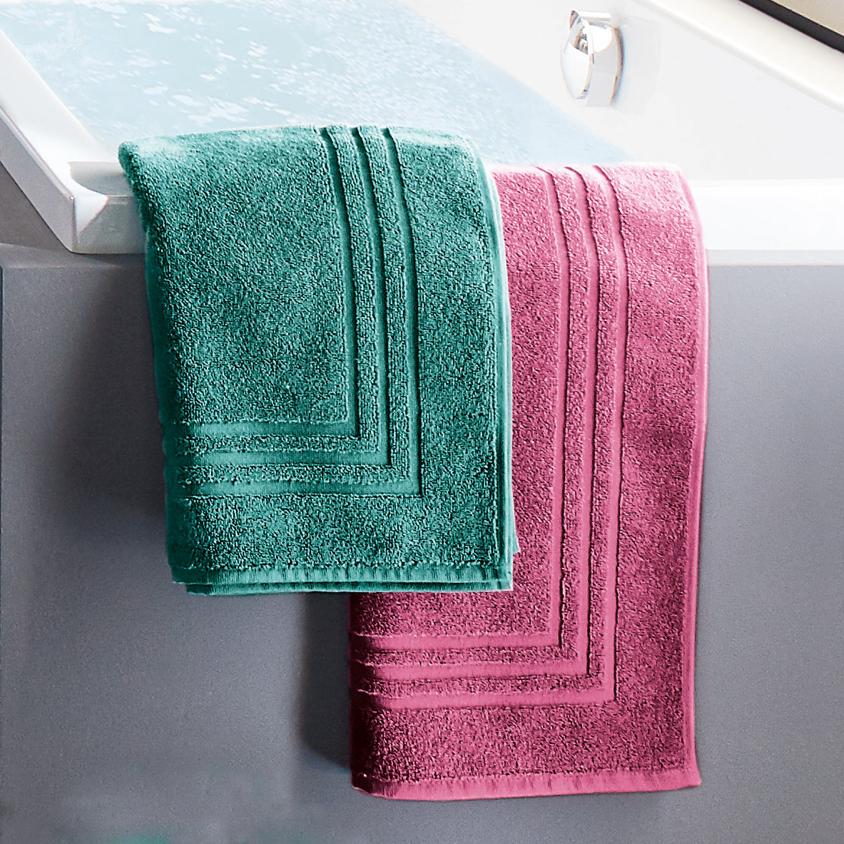 Türkises und pinkes Frottierhandtuch hängen nebeneinander über dem Rand einer Badewanne.