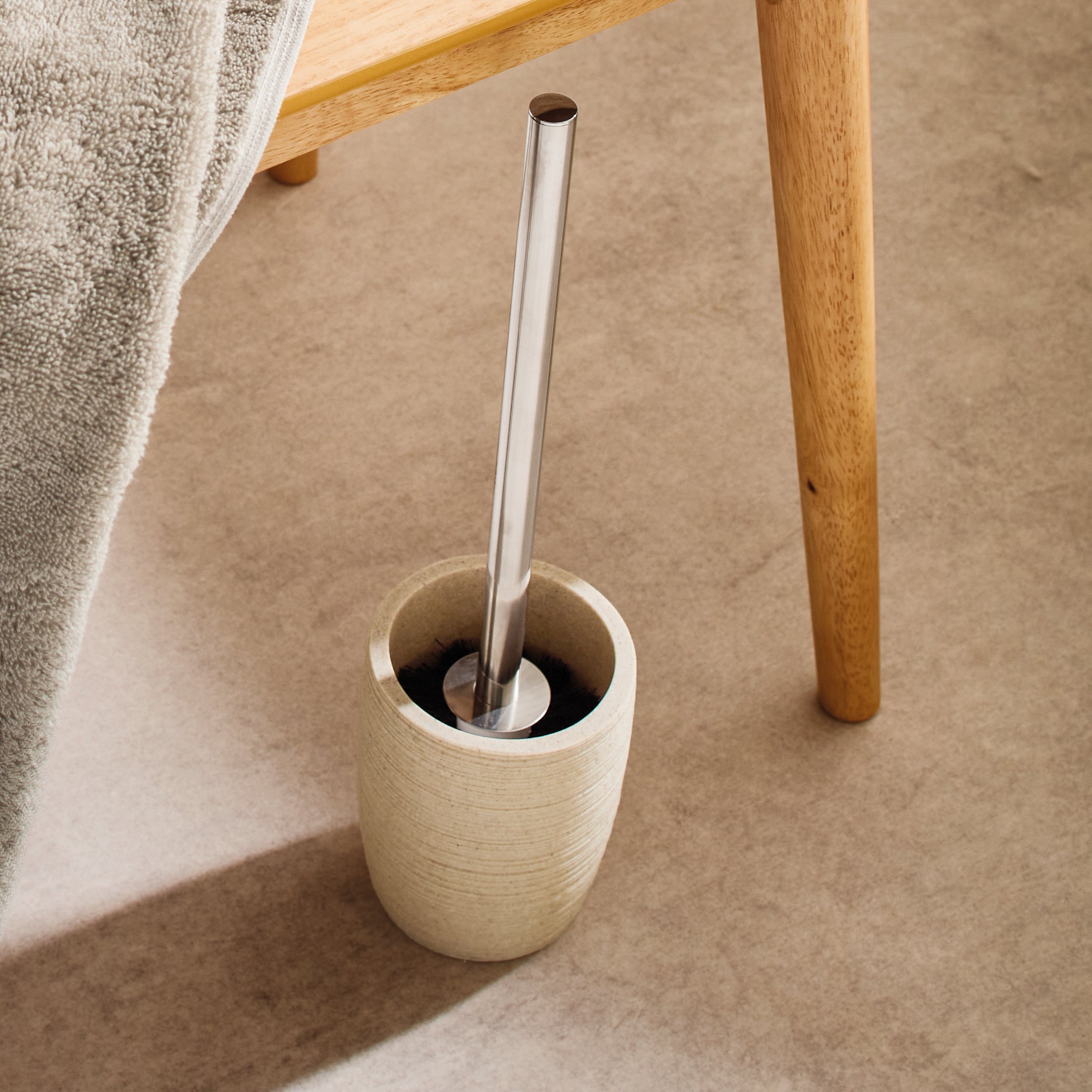 Beige WC-Bürstengarnitur steht neben einem Holzstuhl mit herabhängendem Handtuch.