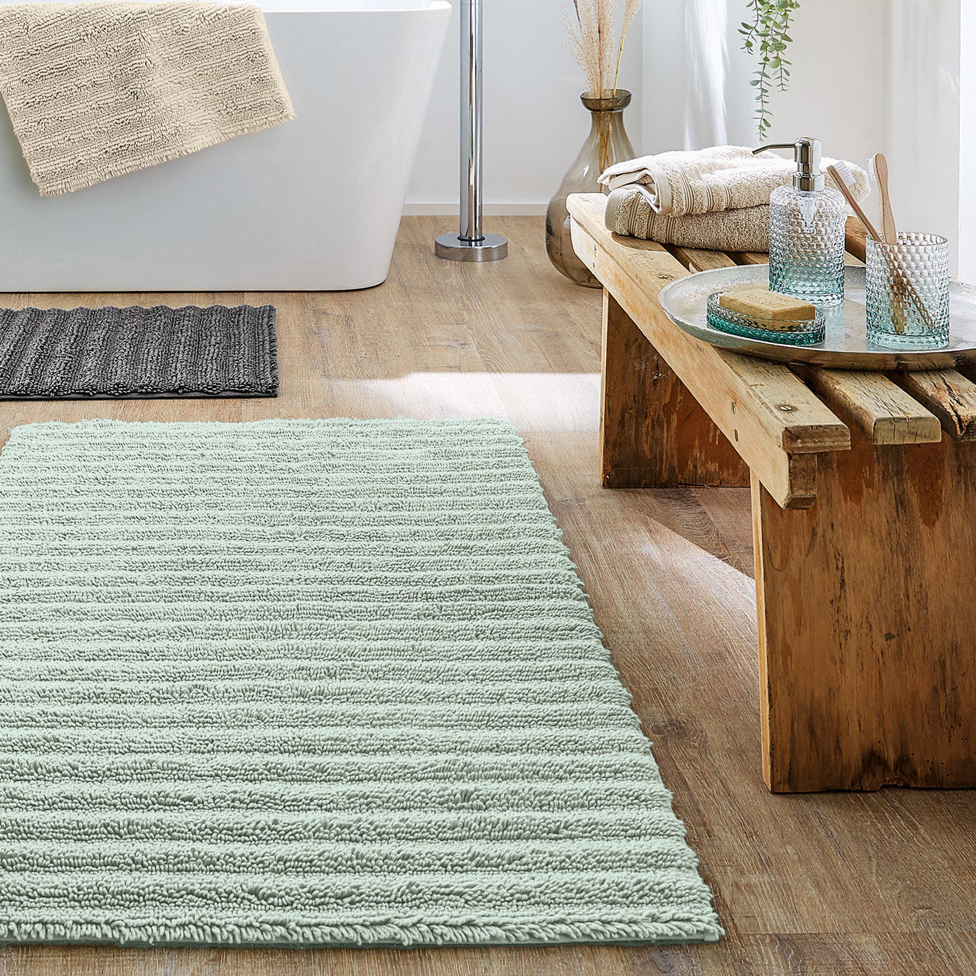 Bad mit grüner Kleine Wolke Eco Collection Badematte und Holzbank mit Handtüchern neben der Badewanne.