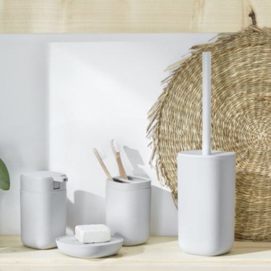 Helle Badaccessoires mit Zahnbürsten, Seife, WC-Bürste und runder Flechtunterlage auf Holz.