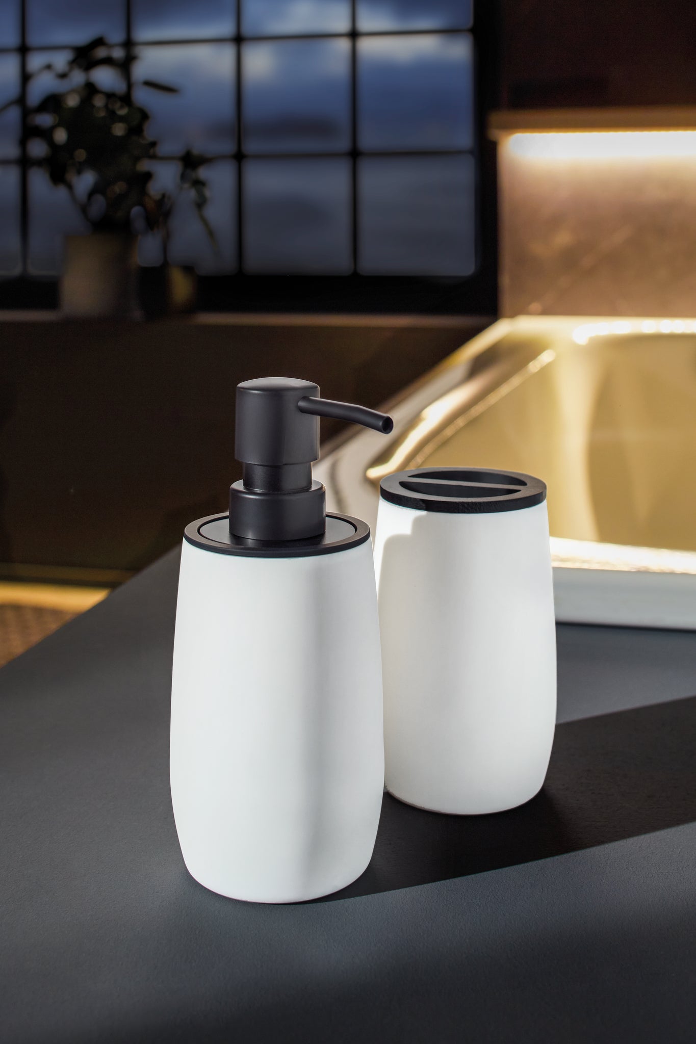 Der Seifenspender DAILY Weiß steht mit passendem Becher auf einem dunklen Waschtisch im Badezimmer.