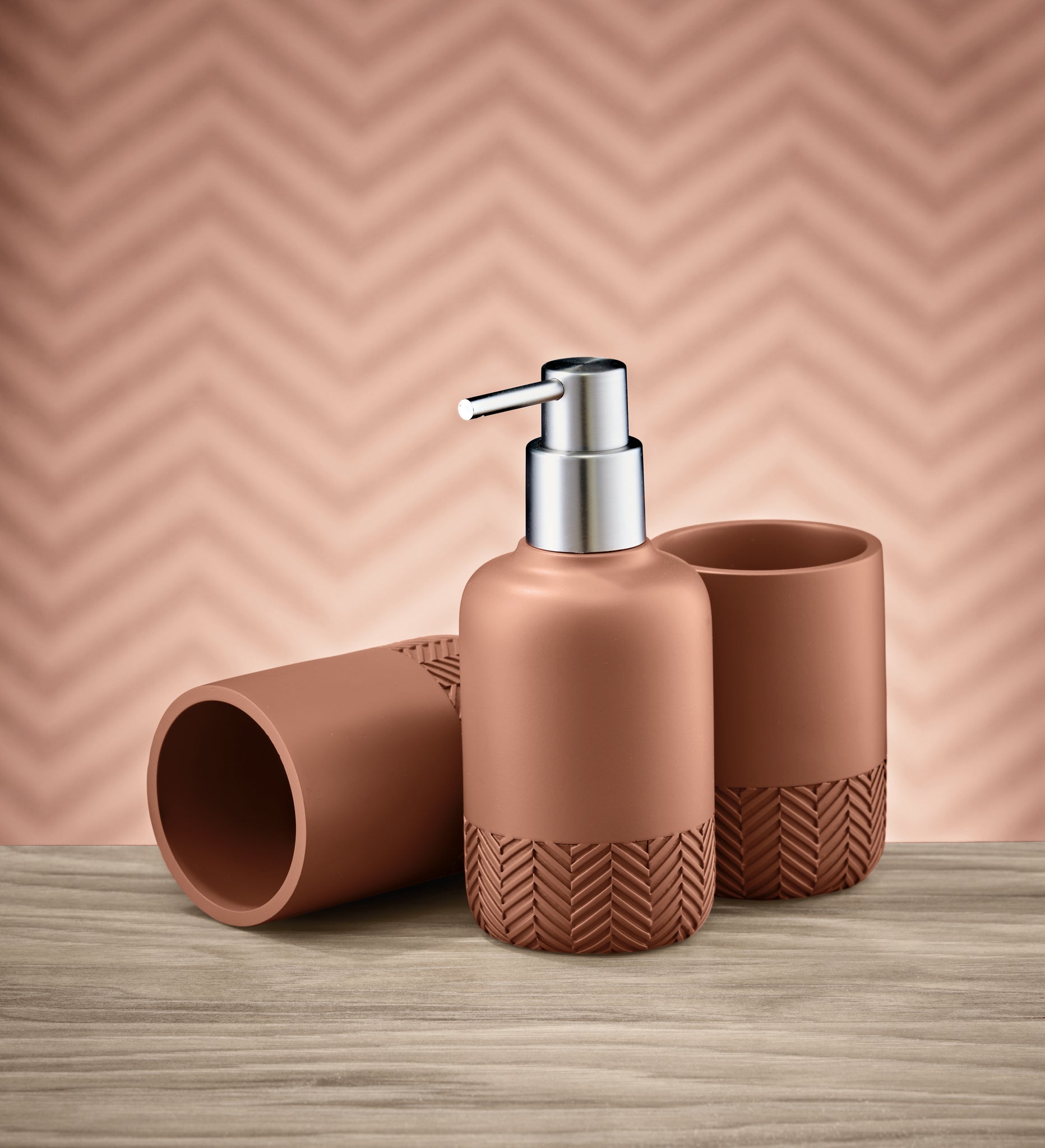 Seifenspender HERRINGBONE Terrabraun mit zwei Bechern auf Holzoberfläche vor gemusterter Wand.