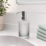 Ein transparenter Glas-Seifenspender mit Chrompumpe steht am Waschbecken.