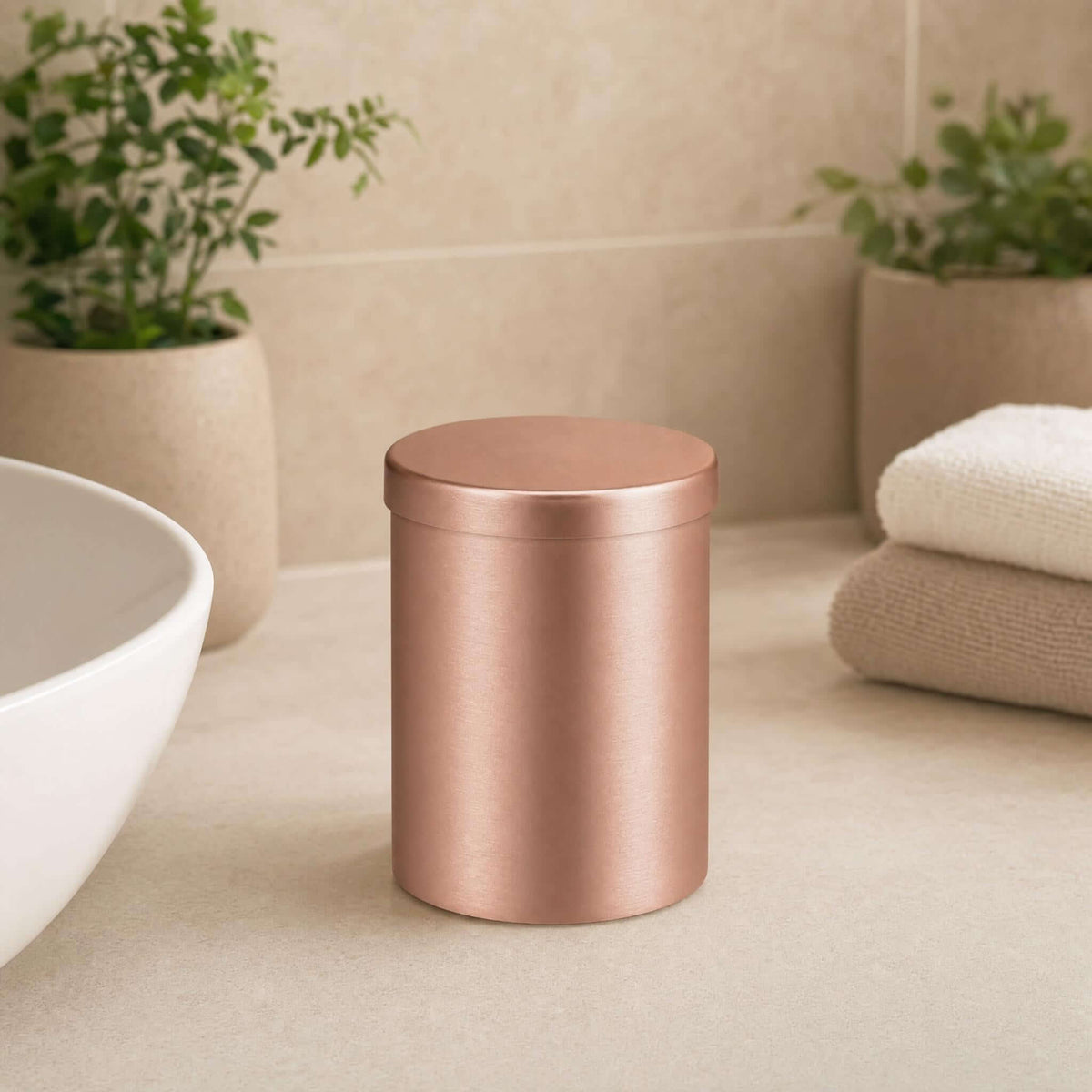Roségoldfarbene Dose im Bad neben Handtüchern, Waschbecken und Pflanzen.