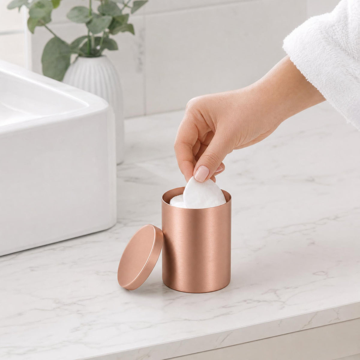 Hand greift im Bad zu Wattepads aus roségoldfarbener Dose auf Marmortheke.