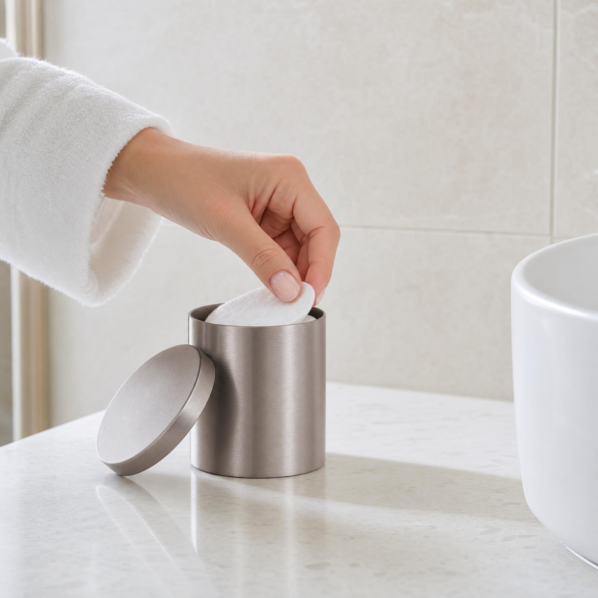 Hand entnimmt Wattepad aus silberfarbener Dose auf hellem Badwaschtisch.