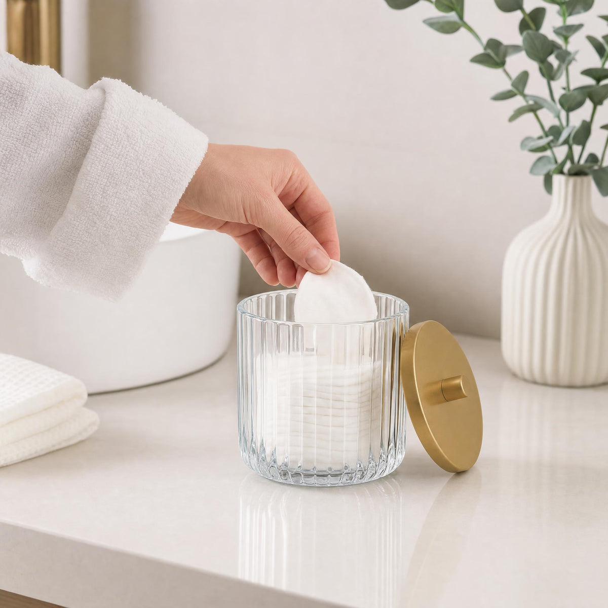Hand nimmt Wattepad aus geriffelter Glasdose im hellen Badezimmer.