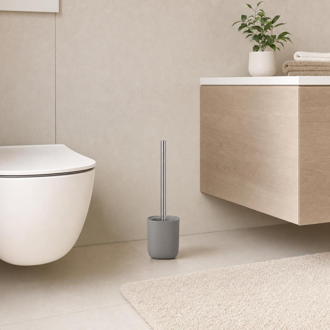 Graue WC-Bürste steht neben Toilette und Holzwaschtisch im Bad.