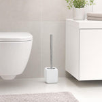 Weiße Toilettenbürste mit Chromgriff steht neben WC und Badschrank.